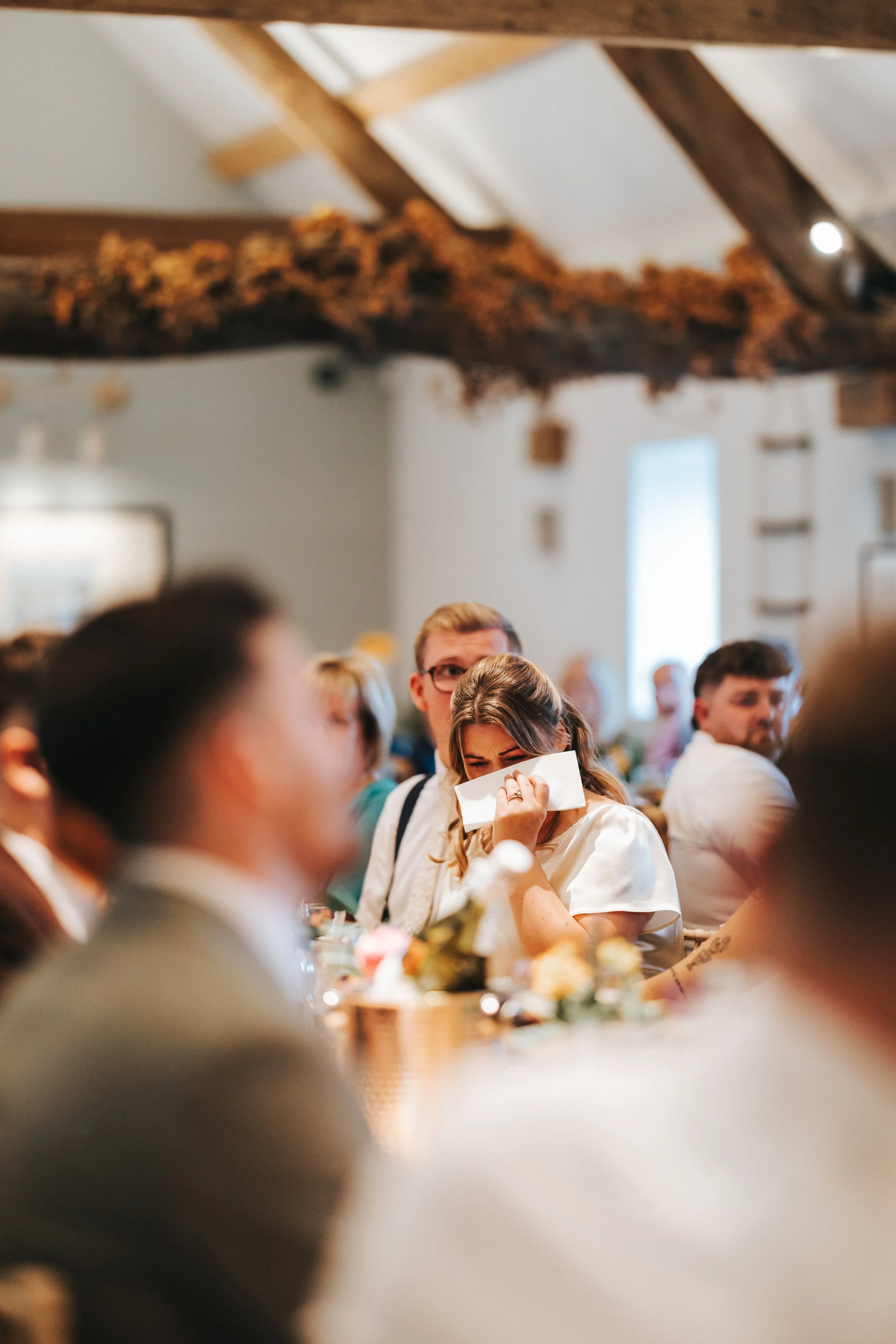 A woman wiping tears with a tissue at a social gathering or event, surrounded by blurred people and decorated table settings in a rustic venue.