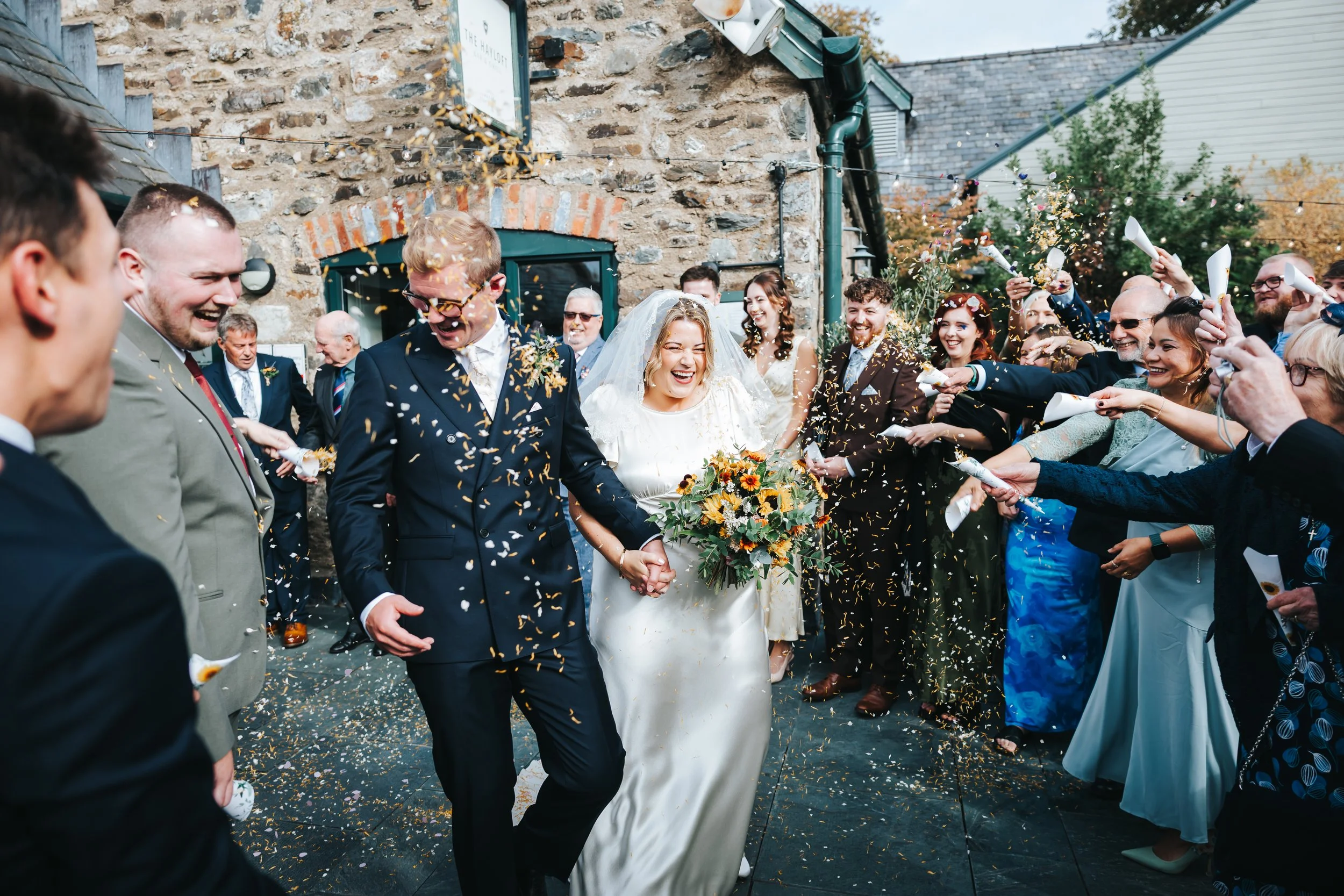 A newlywed couple holding hands and smiling as they walk through a crowd of wedding guests throwing confetti outside a stone building.