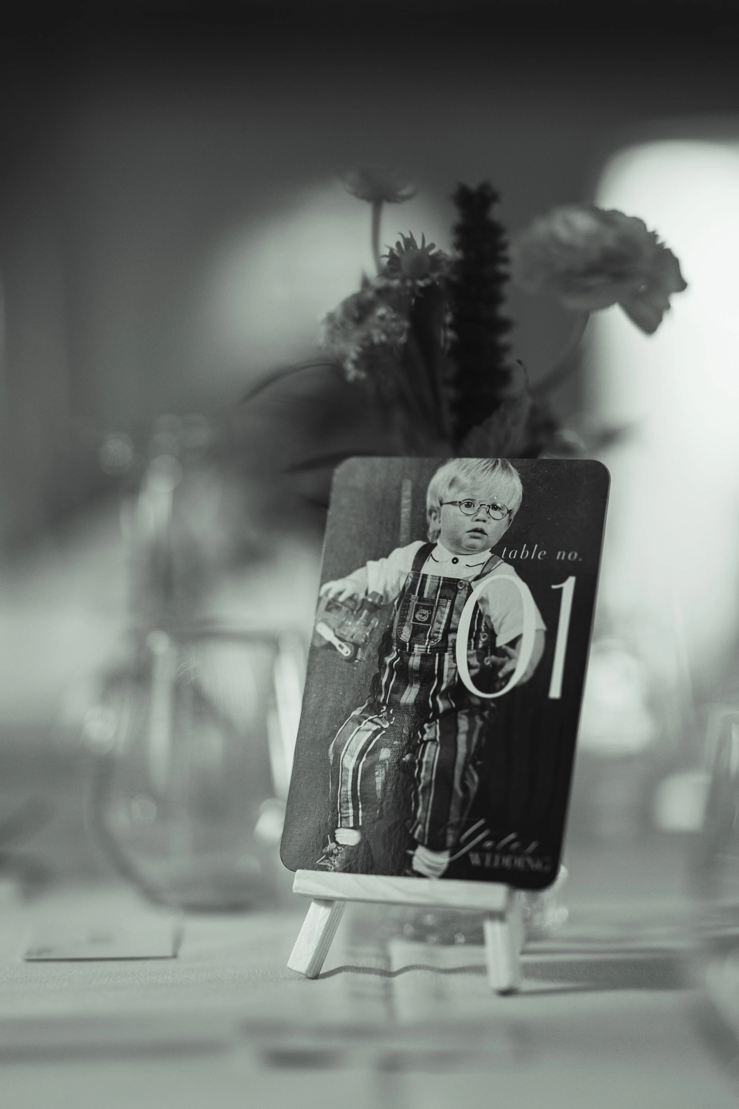 A black and white photo showing a table number card with an image of a young boy wearing glasses and striped overalls, placed on a small easel on a table with flowers and glassware in the background.