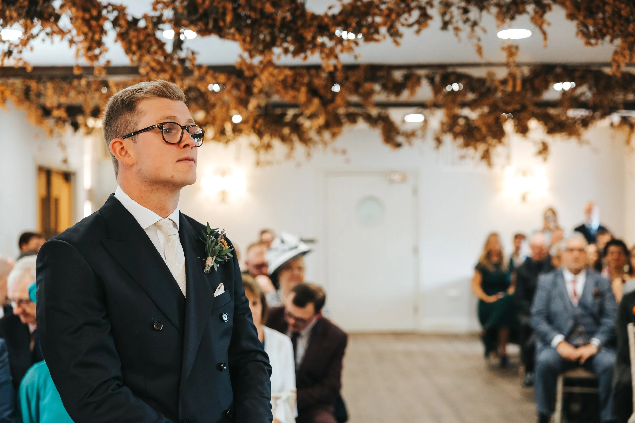 A groom in a dark suit with a white tie and boutonniere stands during a wedding ceremony, with guests seated in the background, in a decorated indoor venue with white walls and brown autumn leaves hanging from the ceiling.