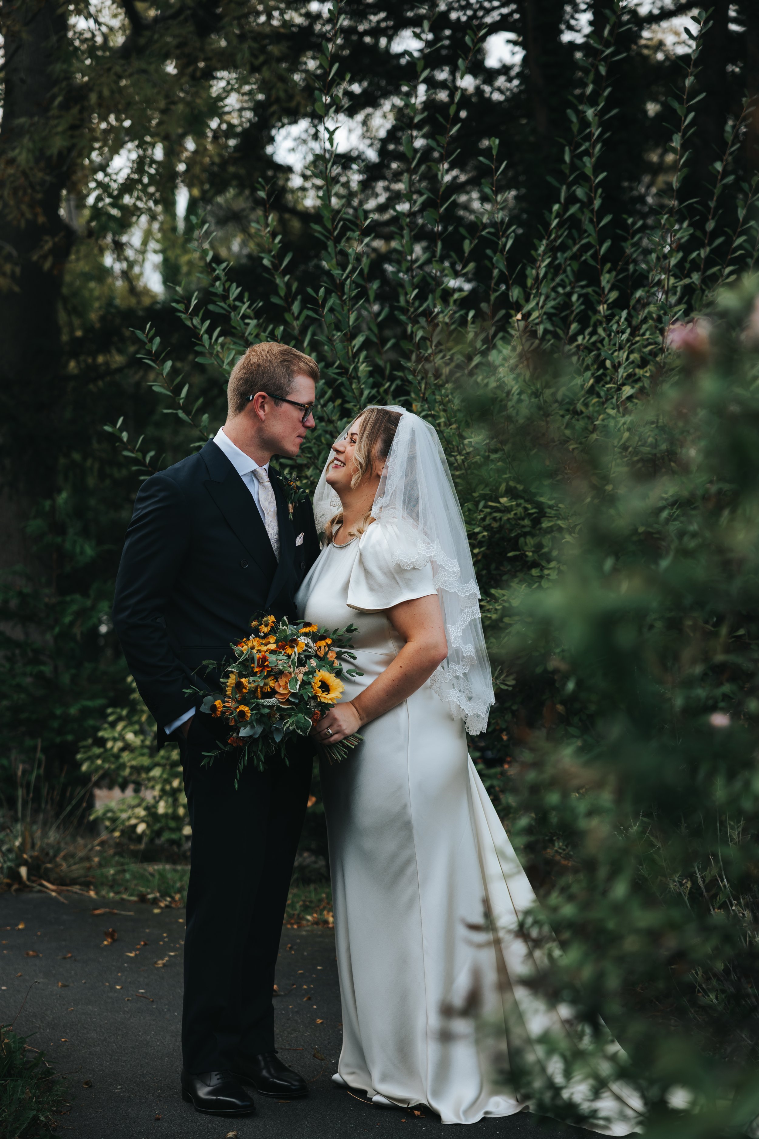 A bride and groom stand close together outdoors, surrounded by greenery, looking lovingly at each other. The bride is holding a bouquet of sunflowers and wearing a white dress with lace veil, while the groom is in a dark suit with glasses.