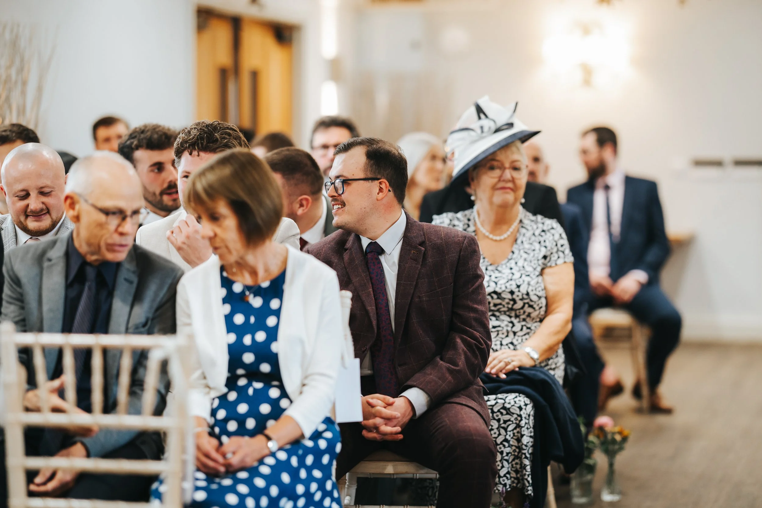 A group of people sitting in a waiting area at a wedding ceremony, dressed in formal attire, including a woman wearing a large white hat with black accents and a pearl necklace.