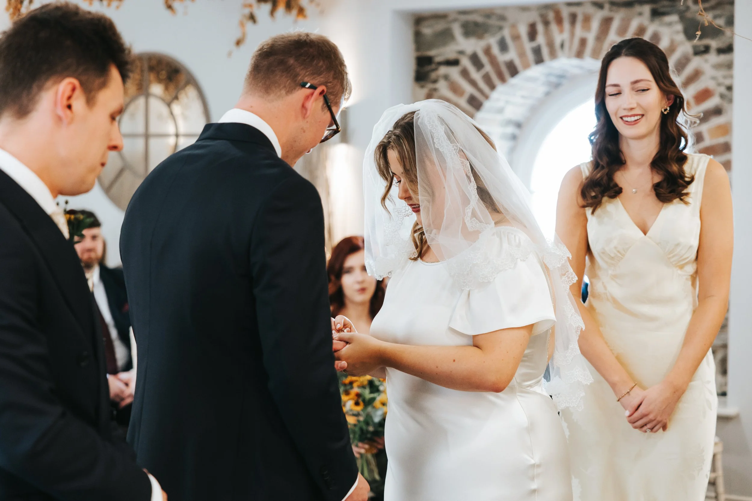 A bride and groom exchange rings during a wedding ceremony while a woman in a cream dress looks on smiling inside a rustic venue with brick walls and round windows.