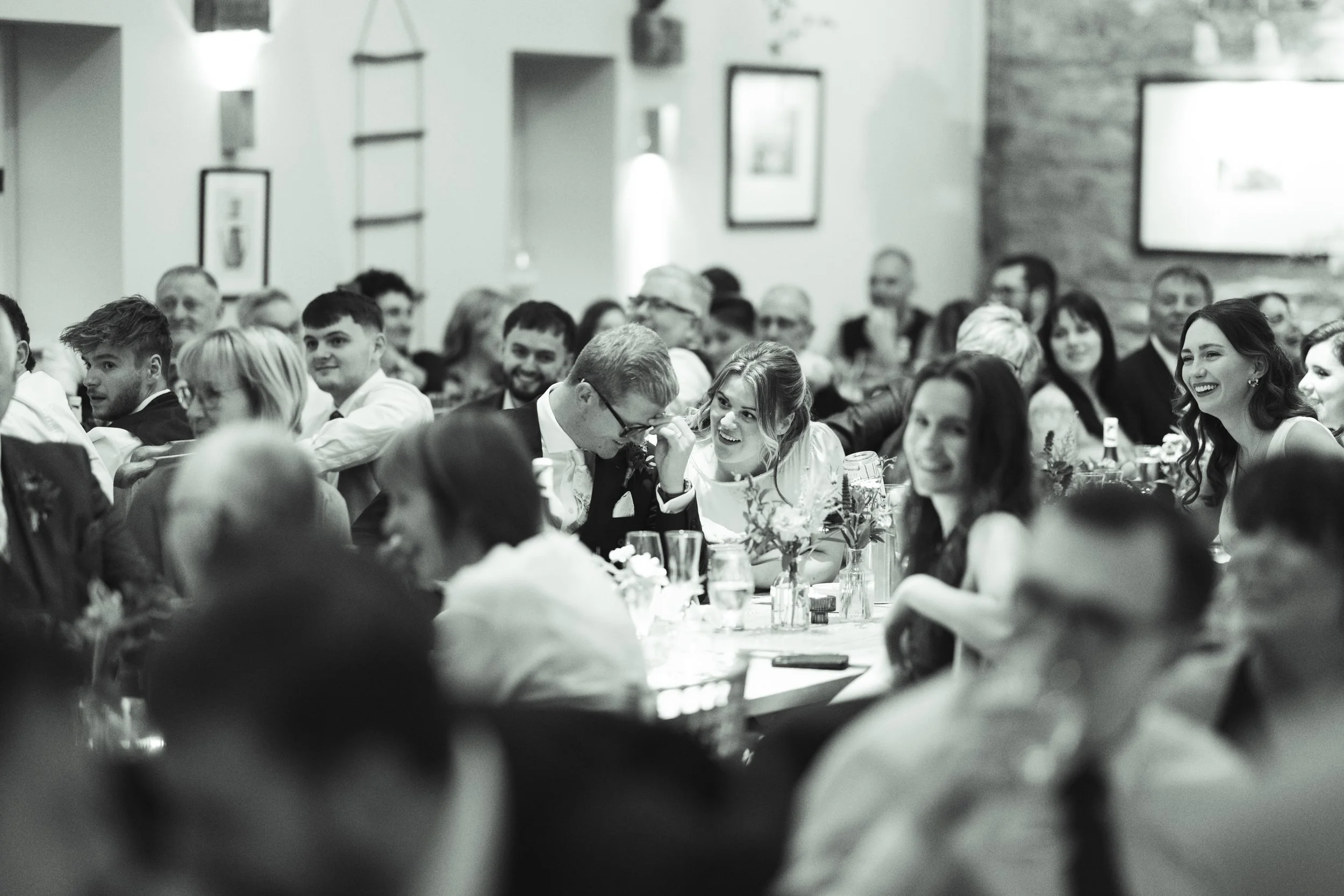People gathered at a formal event, sitting at tables with floral centerpieces, smiling and engaging in conversations, in a decorated indoor venue.
