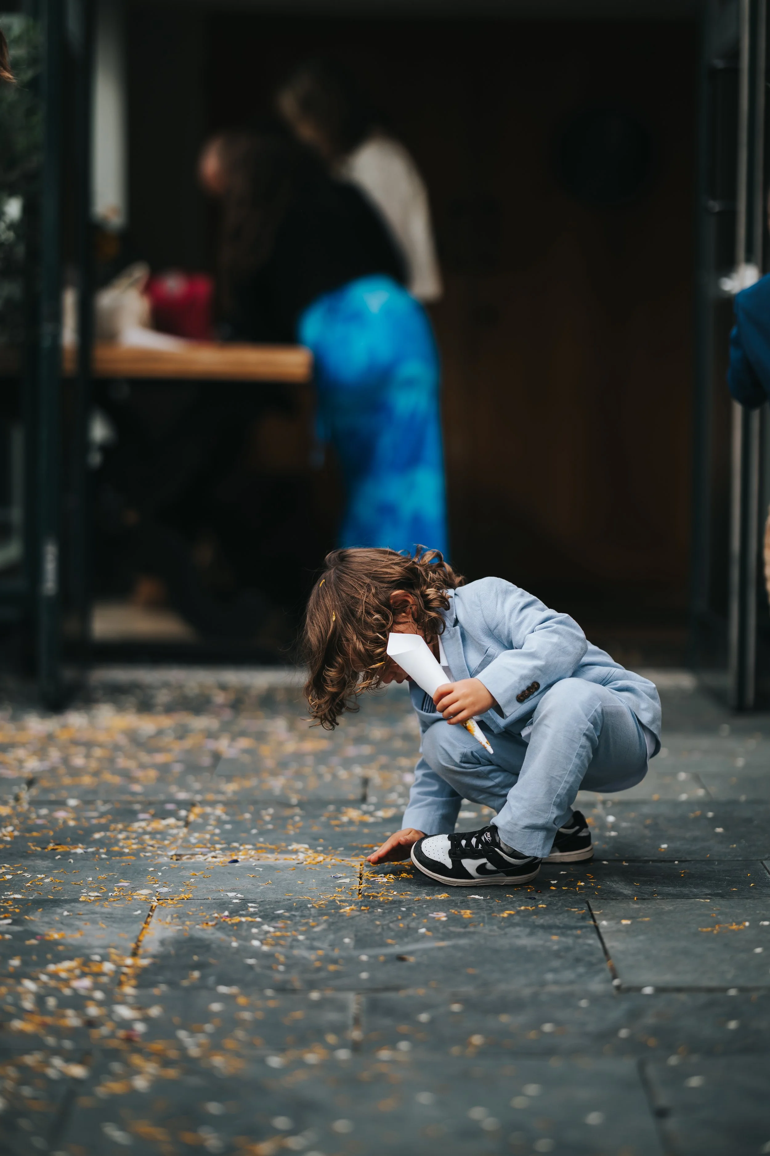 Child in a gray suit squatting outdoors, picking up and examining scattered yellow and white flower petals or confetti on the ground, holding a white plastic bottle.