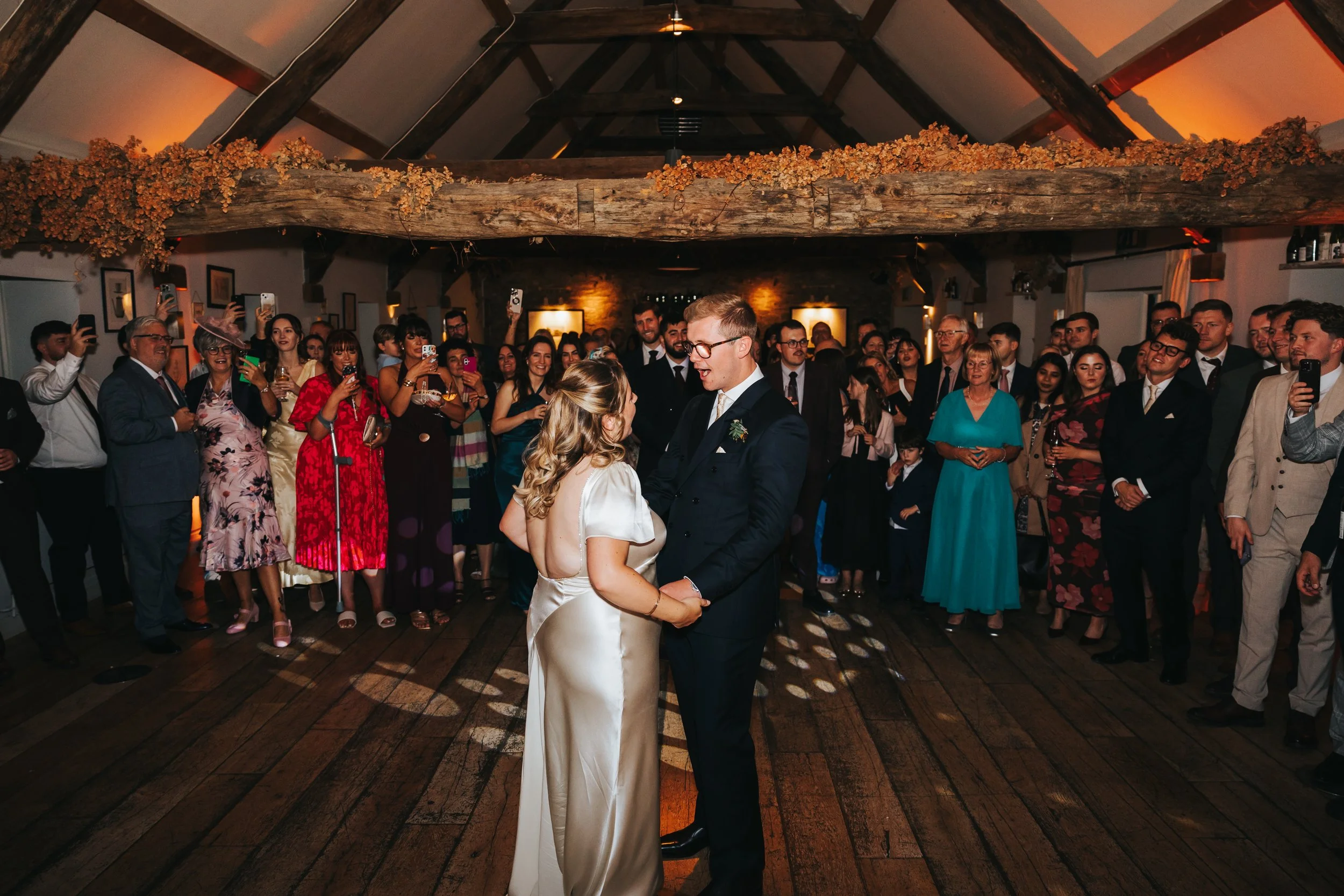 A bride and groom dance in center of reception hall, surrounded by guests watching and taking photos at wedding celebration.