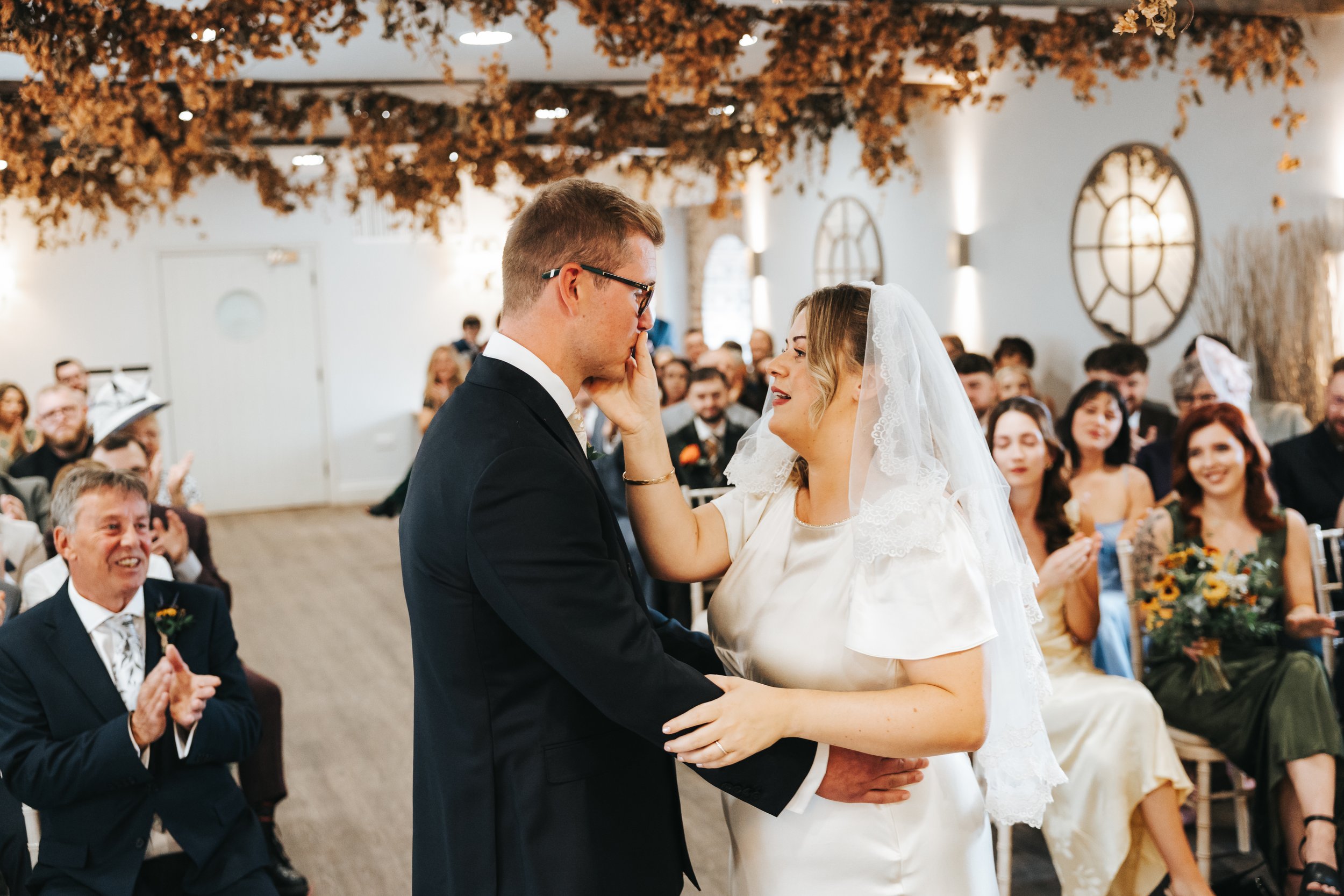 A bride and groom exchange vows during their wedding ceremony in a decorated indoor venue with guests clapping and smiling in the background.