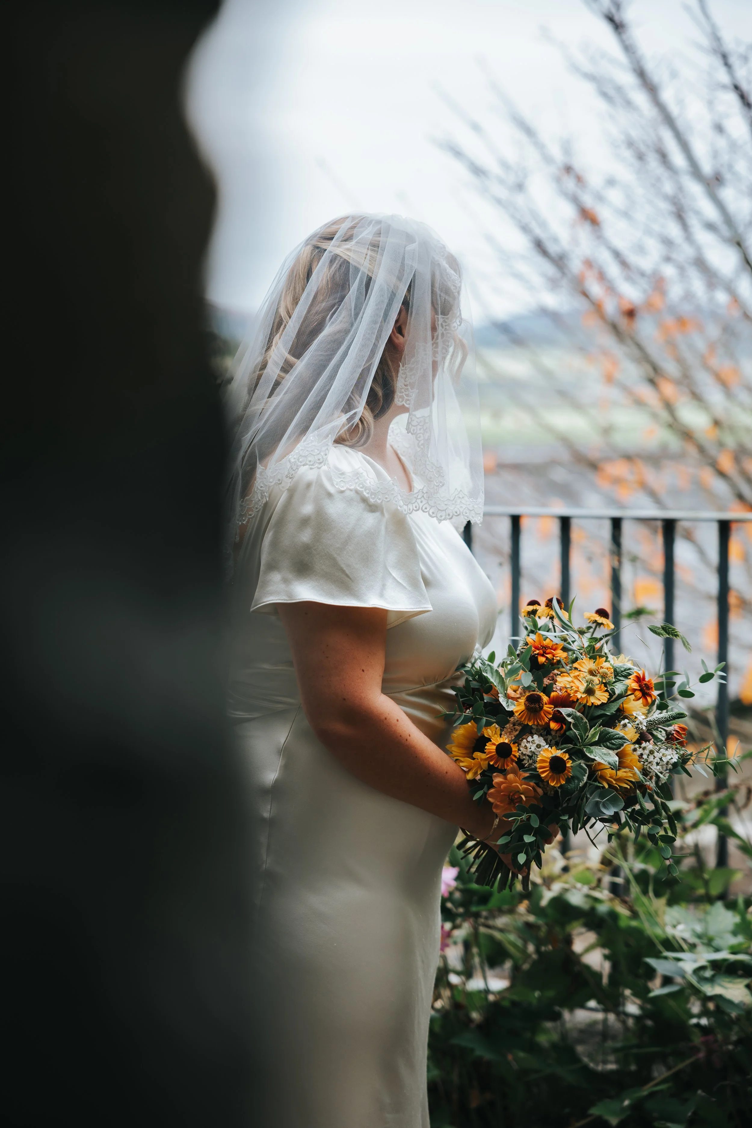 A bride wearing a white satin dress and a lace veil holds a colorful bouquet of sunflowers and other flowers outdoors near a black metal railing.