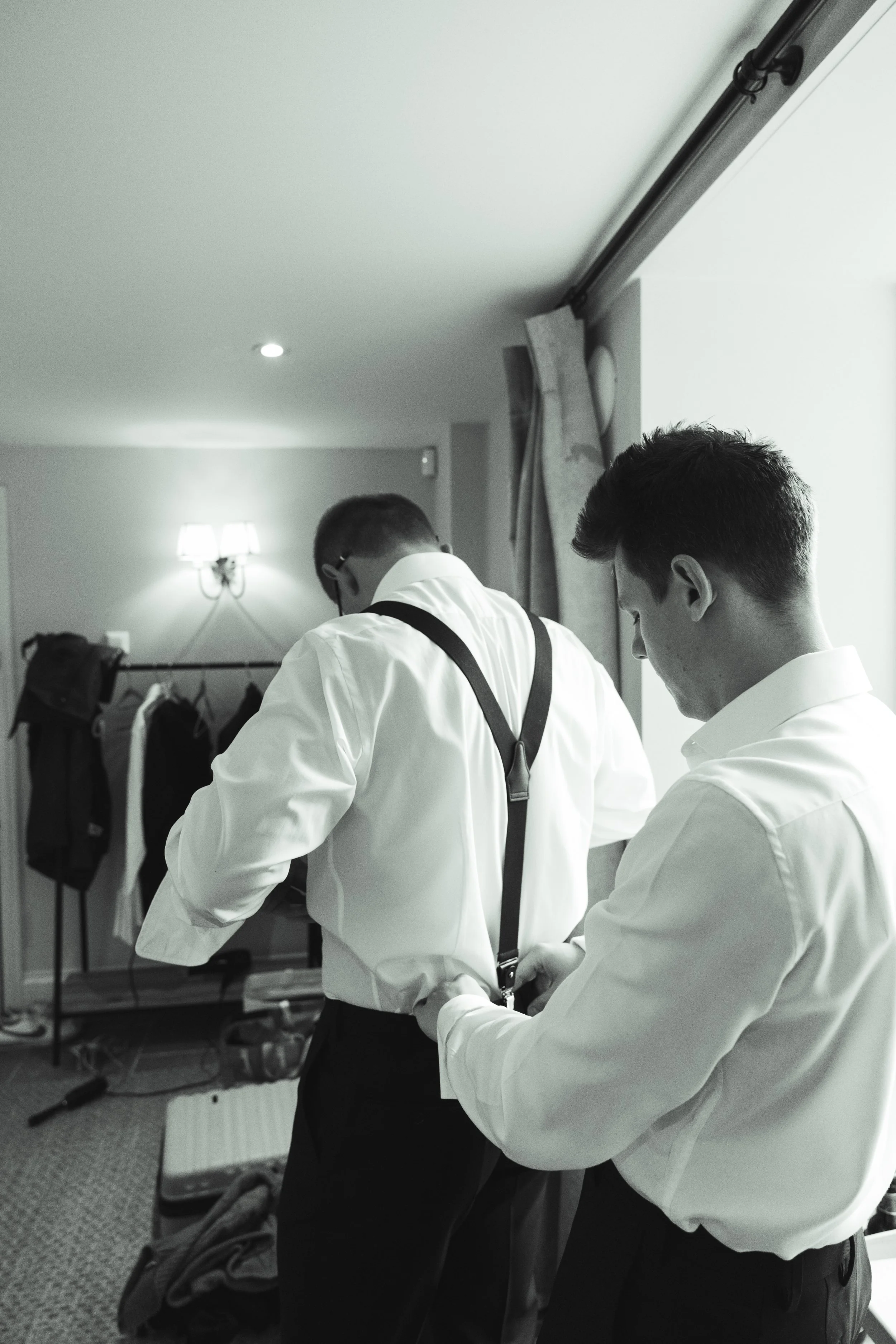 Two men in white shirts and black suspenders adjusting their belts in a hotel room.