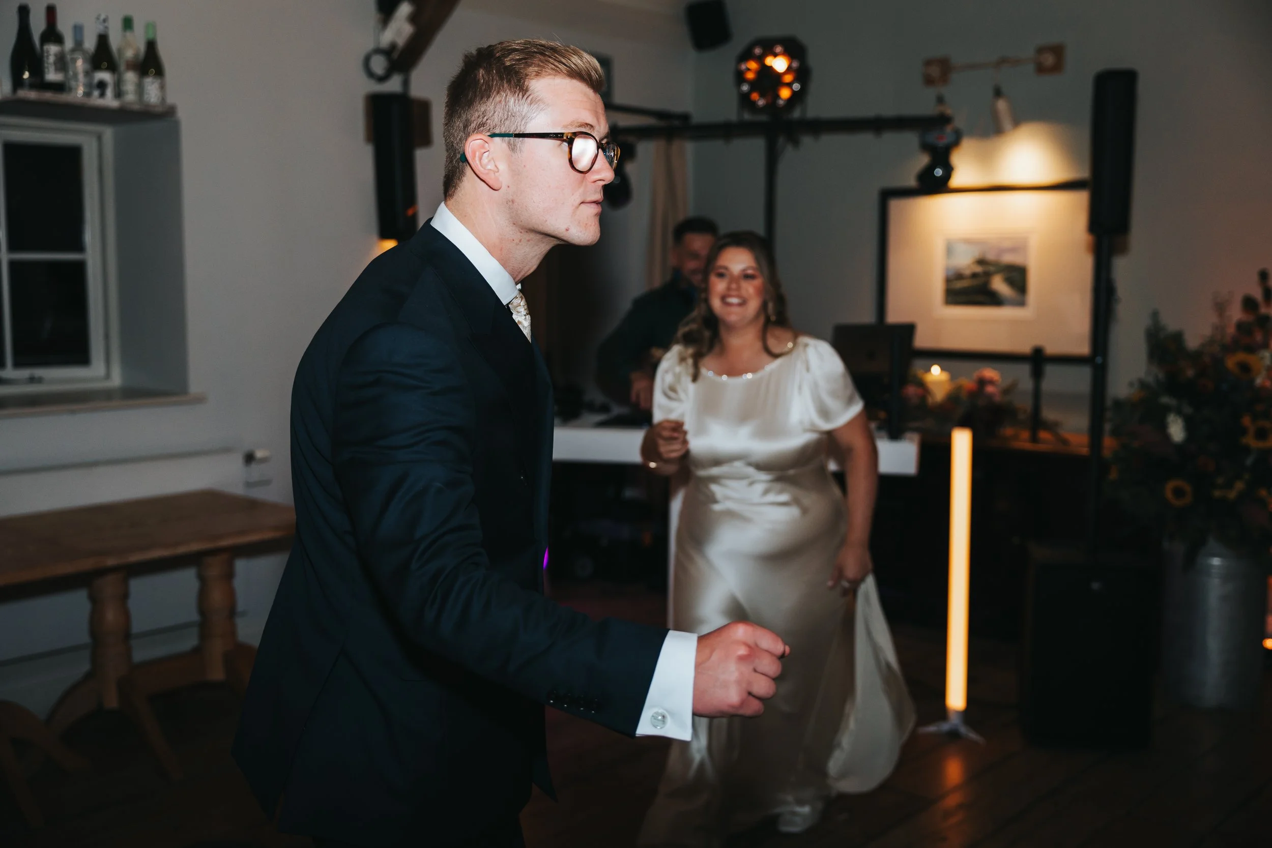 A man in a black suit and glasses is dancing at a wedding reception, with a smiling woman in a white dress in the background.