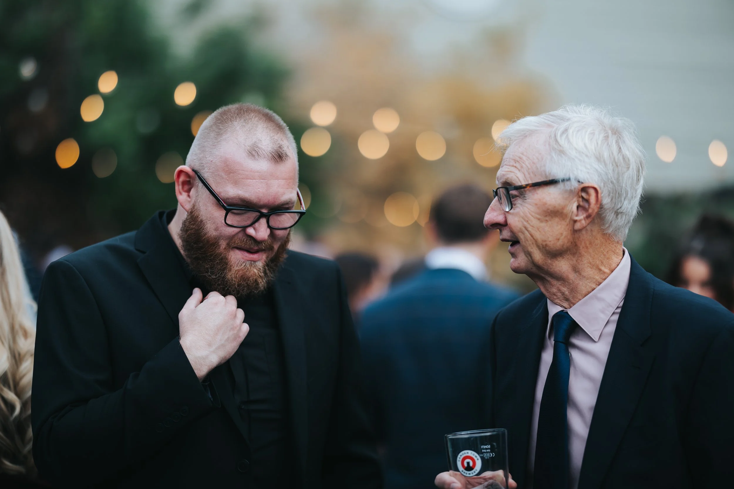 Two men, one with a beard and glasses, the other elderly with white hair and glasses, are talking at a social event, with blurry string lights in the background.