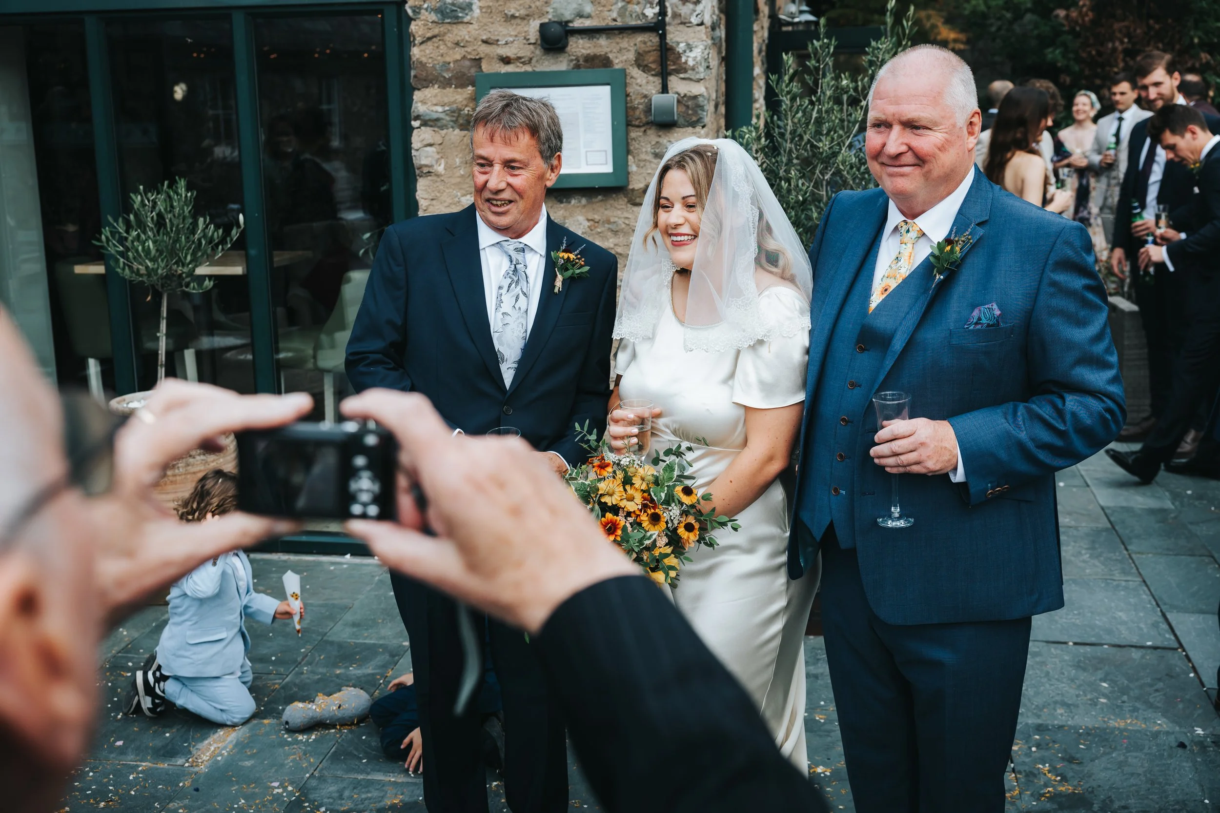 A bride in a white wedding dress and veil is smiling while holding a bouquet of flowers, standing between two men in suits, at an outdoor wedding reception, with guests in the background.