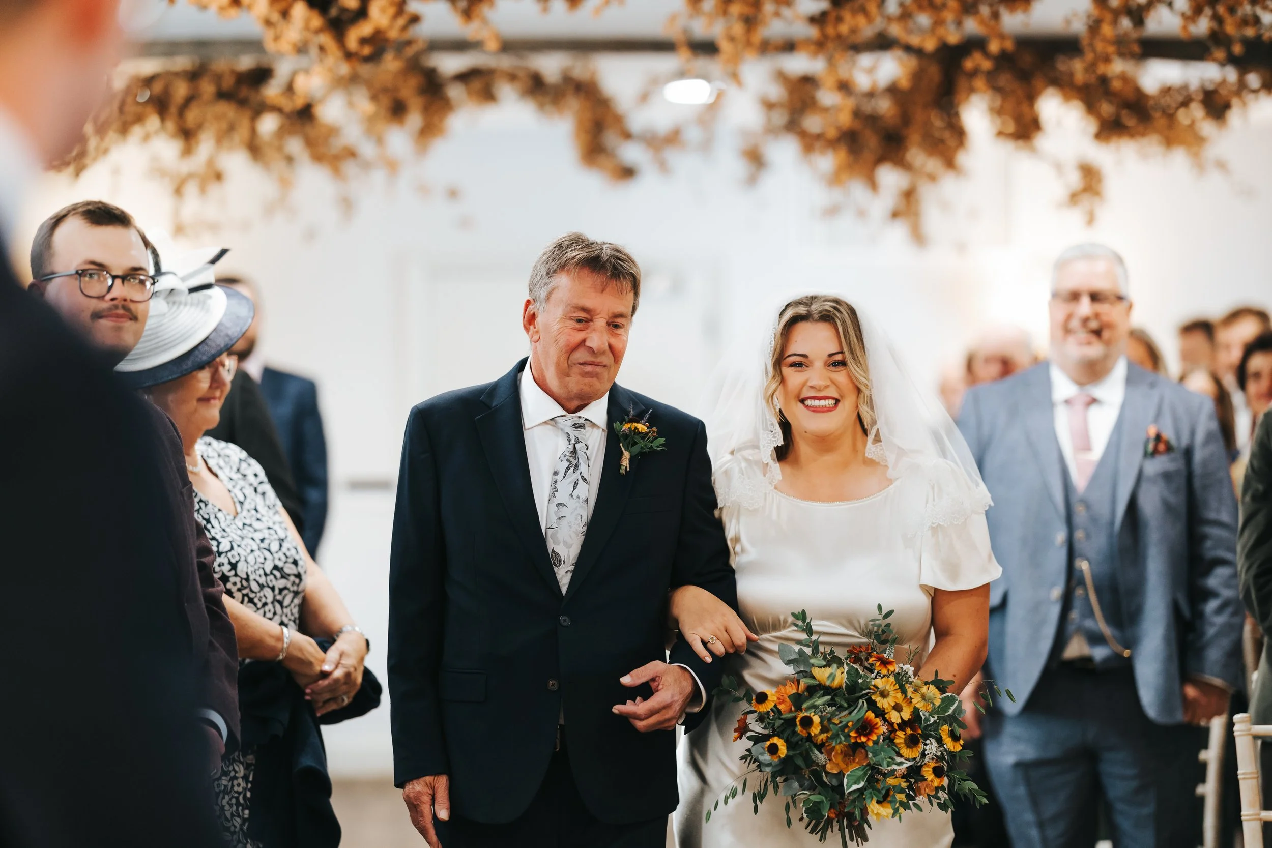 Bride walking down the aisle holding a bouquet of flowers, accompanied by an elderly man, at a wedding ceremony with guests in the background. The bride is smiling and wearing a white dress and veil. The elderly man is dressed in a dark suit with a b