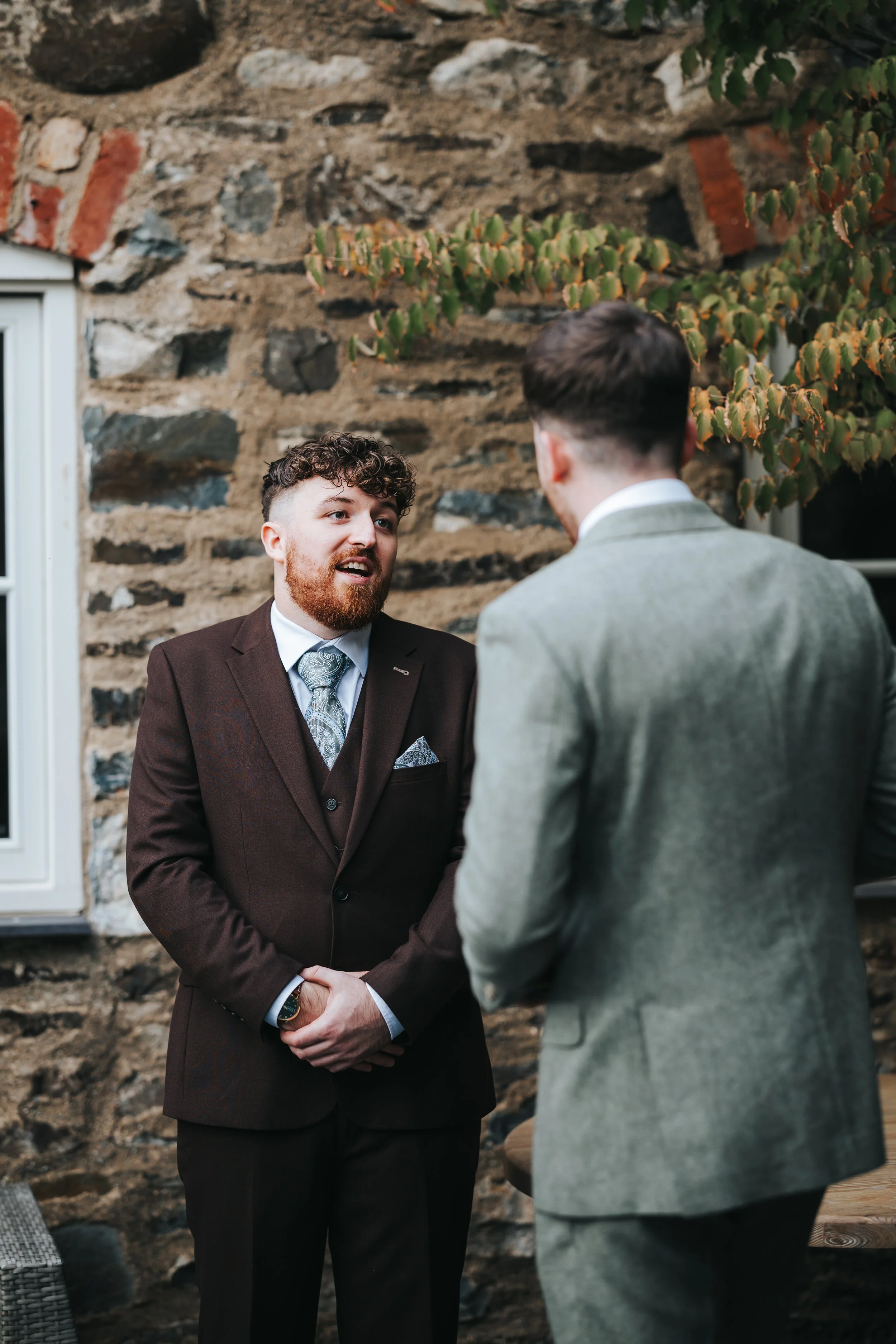 Two men in suits having a conversation outdoors against a stone wall with vine leaves.