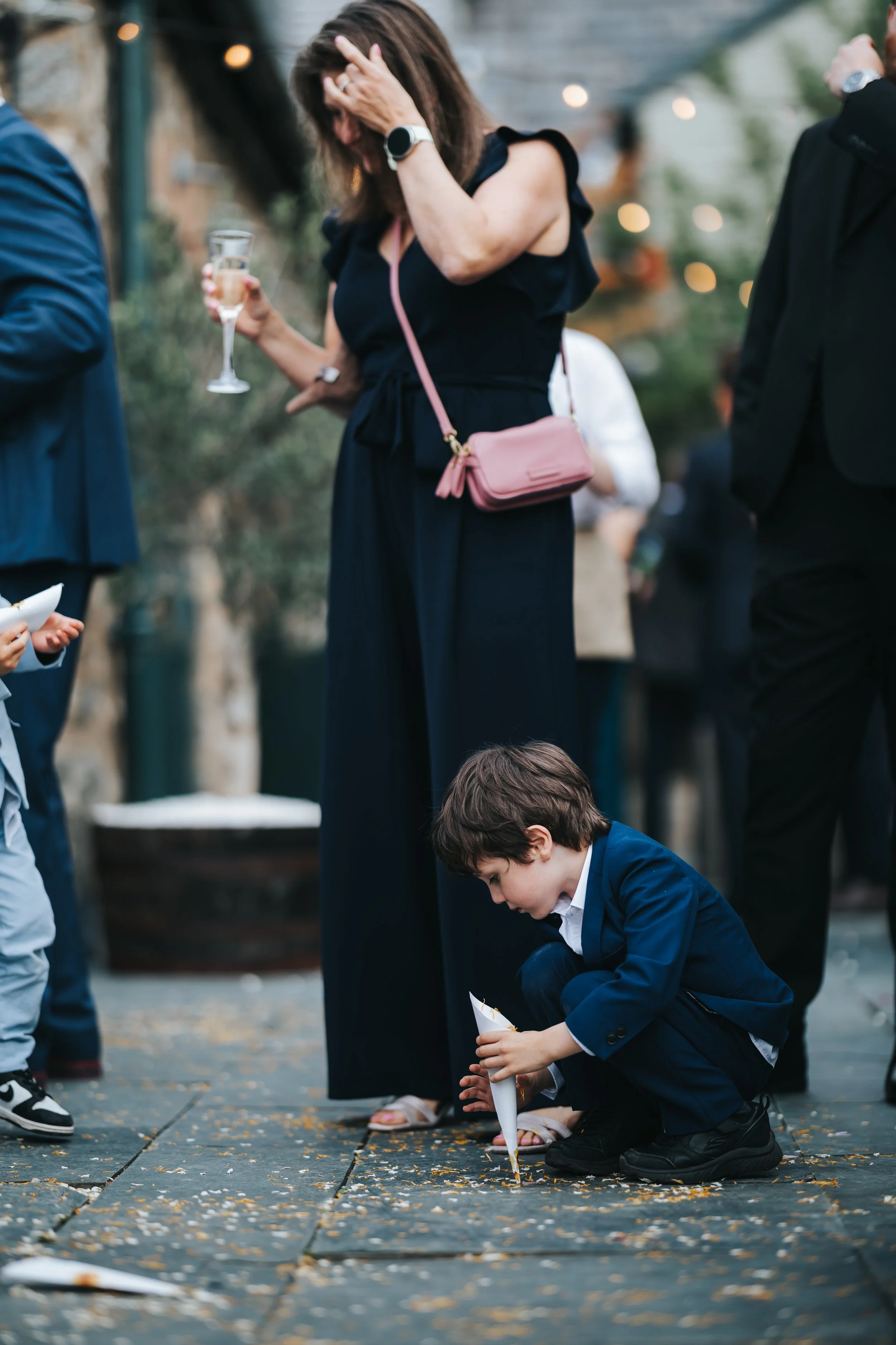 A young boy in a navy suit is crouched on the ground, holding a paper cone, likely filled with confetti or rice, during a celebratory outdoor event. Behind him, a woman in a dark dress with a pink purse and a smartwatch is wiping her face, holding a 