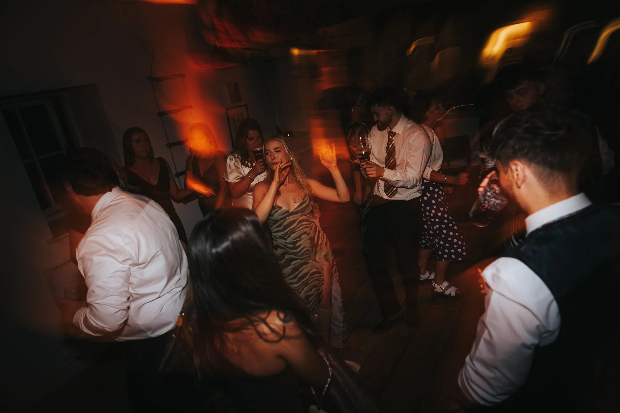 People dancing and socializing at a party in a dimly lit room with orange and yellow lights.