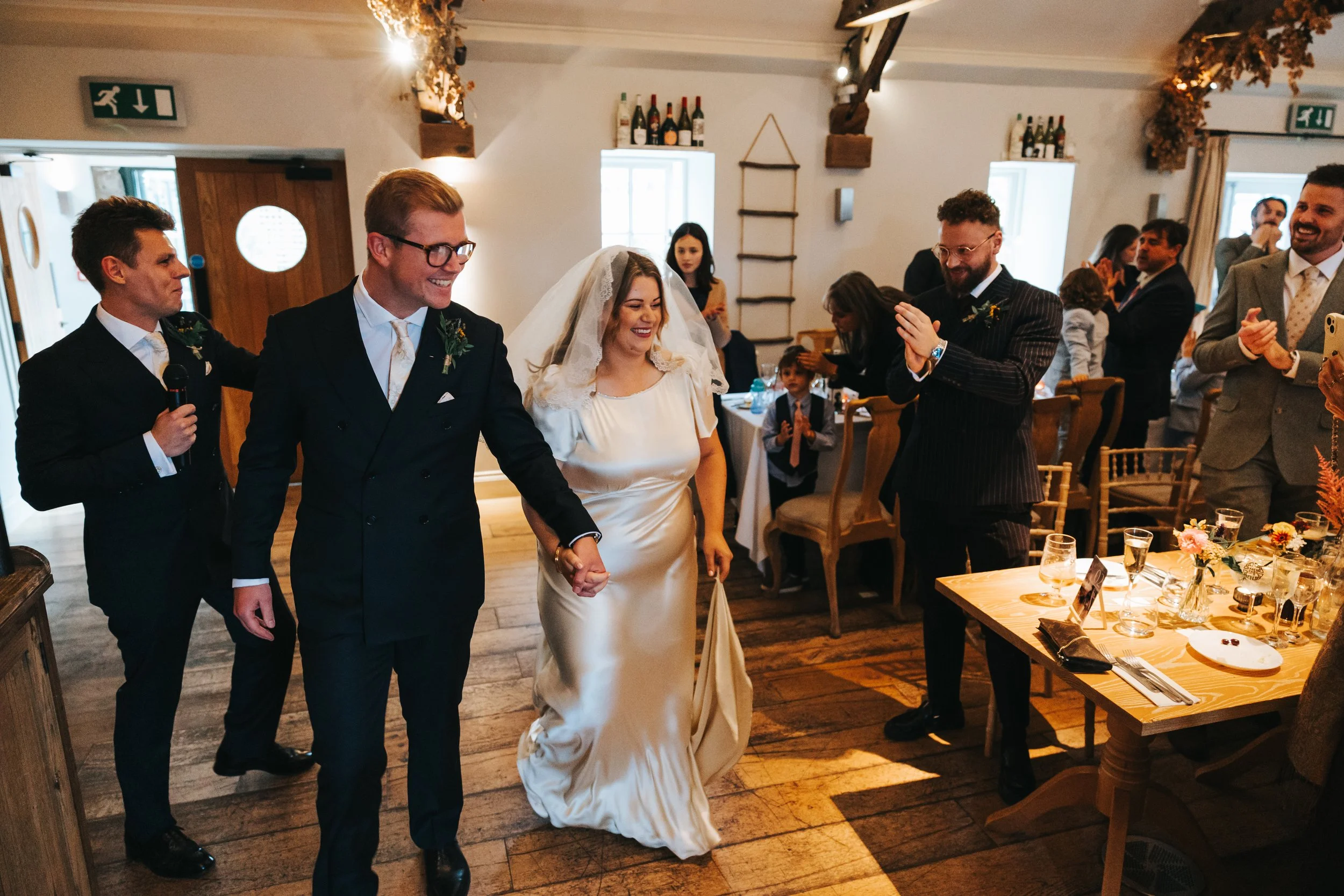 A bride and groom holding hands and smiling as they walk into a wedding reception, surrounded by guests clapping and celebrating in a cozy, decorated dining area.