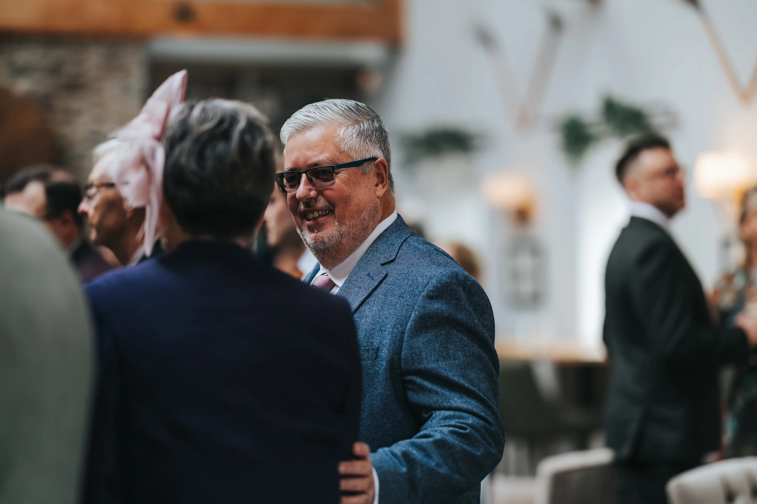 People at a social event, dressed formally, with a man smiling in the foreground, engaged in conversation in a well-lit, decorated indoor setting.