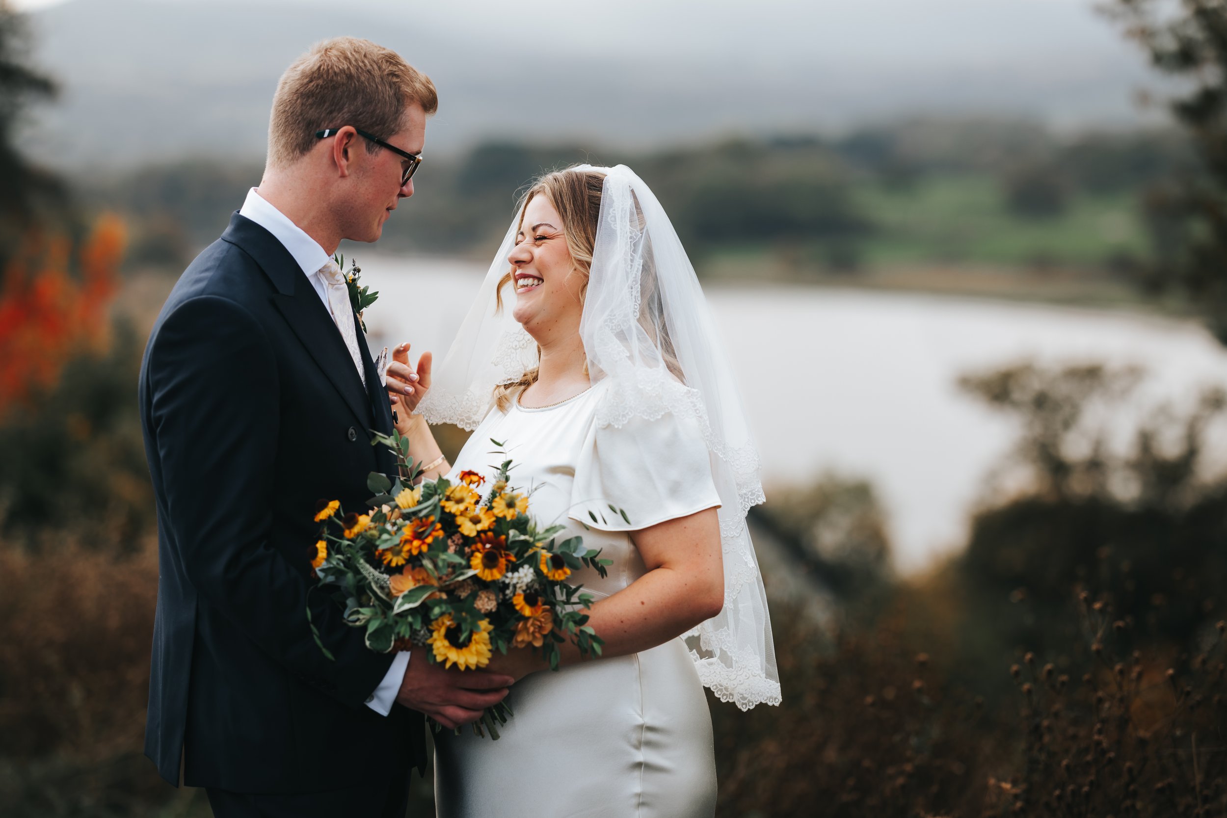 A bride and groom share a joyful moment outdoors during their wedding. The bride is smiling and wearing a white dress with a lace veil, holding a bouquet of sunflowers and greenery. The groom is in a dark suit, glasses, and is smiling back at her. Th