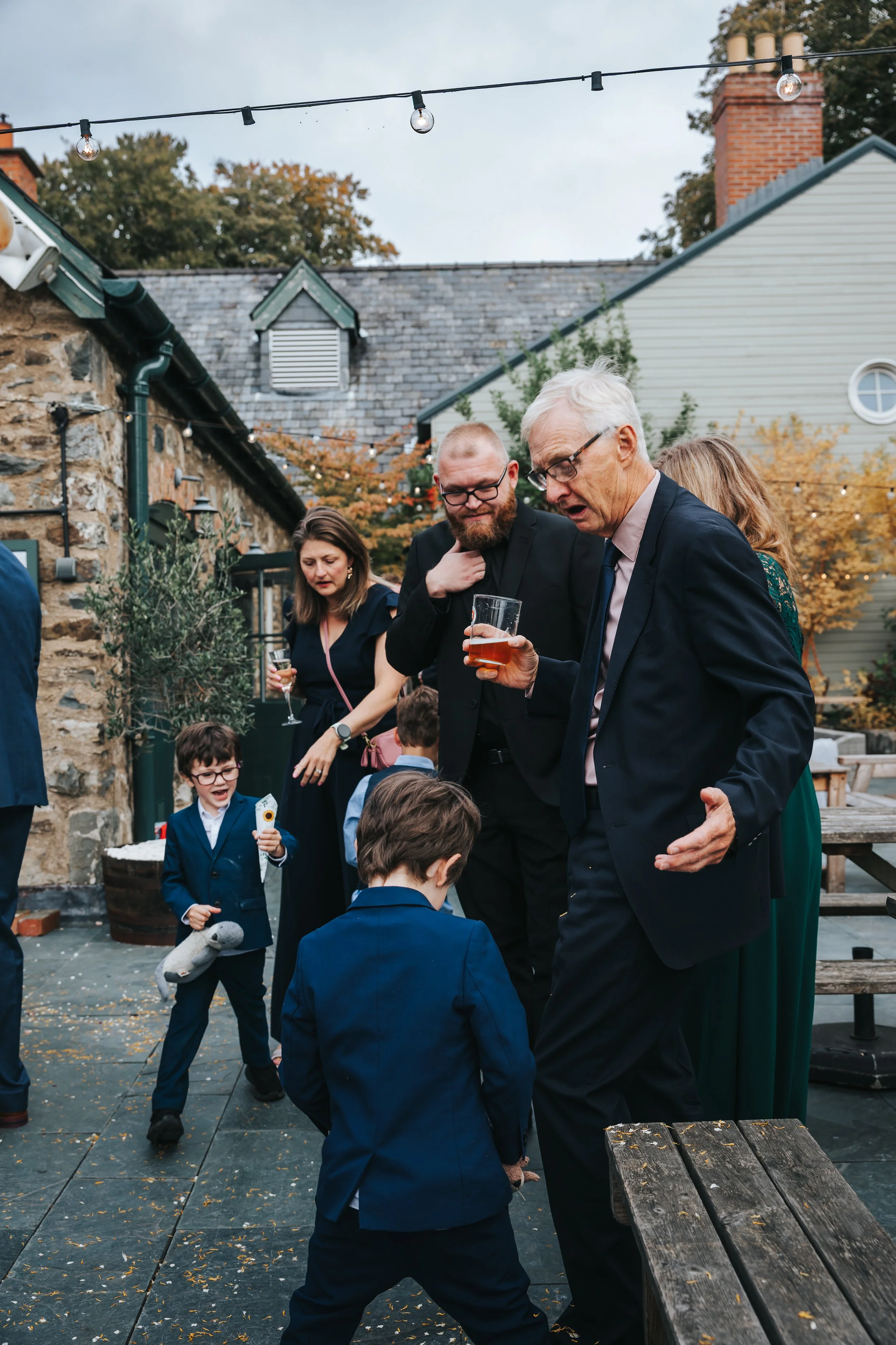 Adults and children at an outdoor gathering, with some adults holding drinks, in front of buildings with autumn trees and string lights overhead.