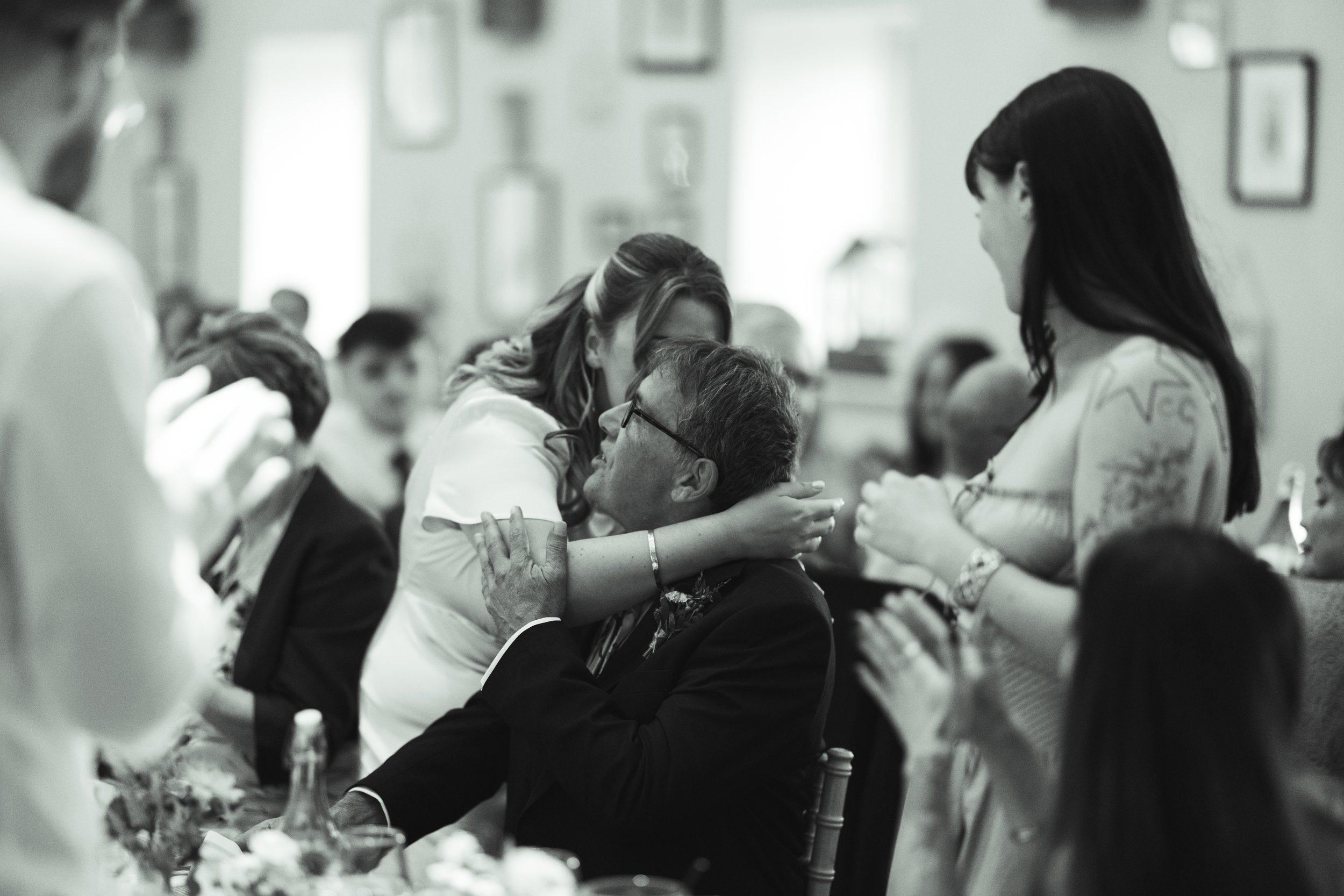 A woman is hugging an older man at a gathering or celebration, surrounded by other people clapping and smiling.