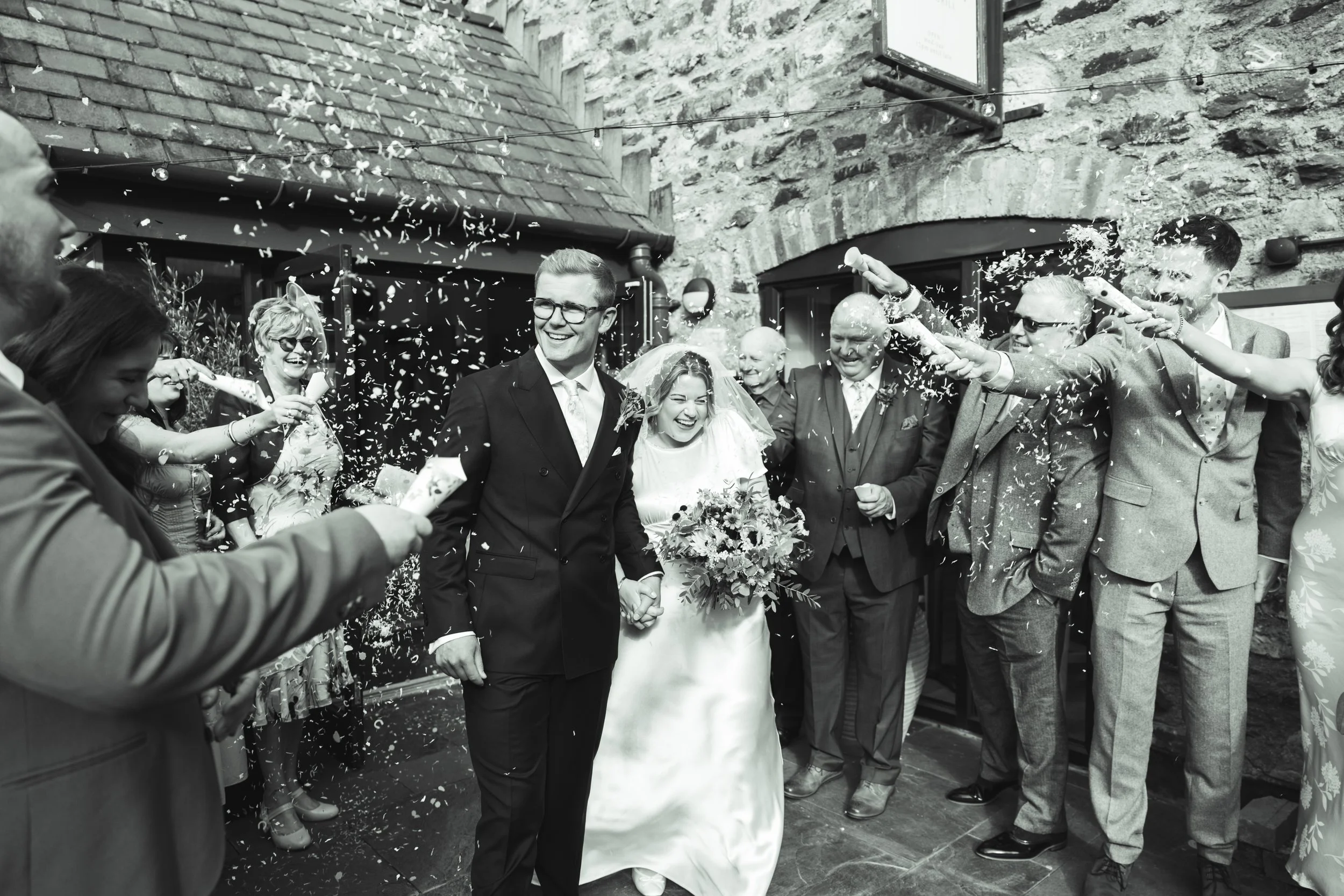 A wedding celebration with a bride and groom holding hands and smiling, surrounded by friends and family throwing confetti, outside a stone building.