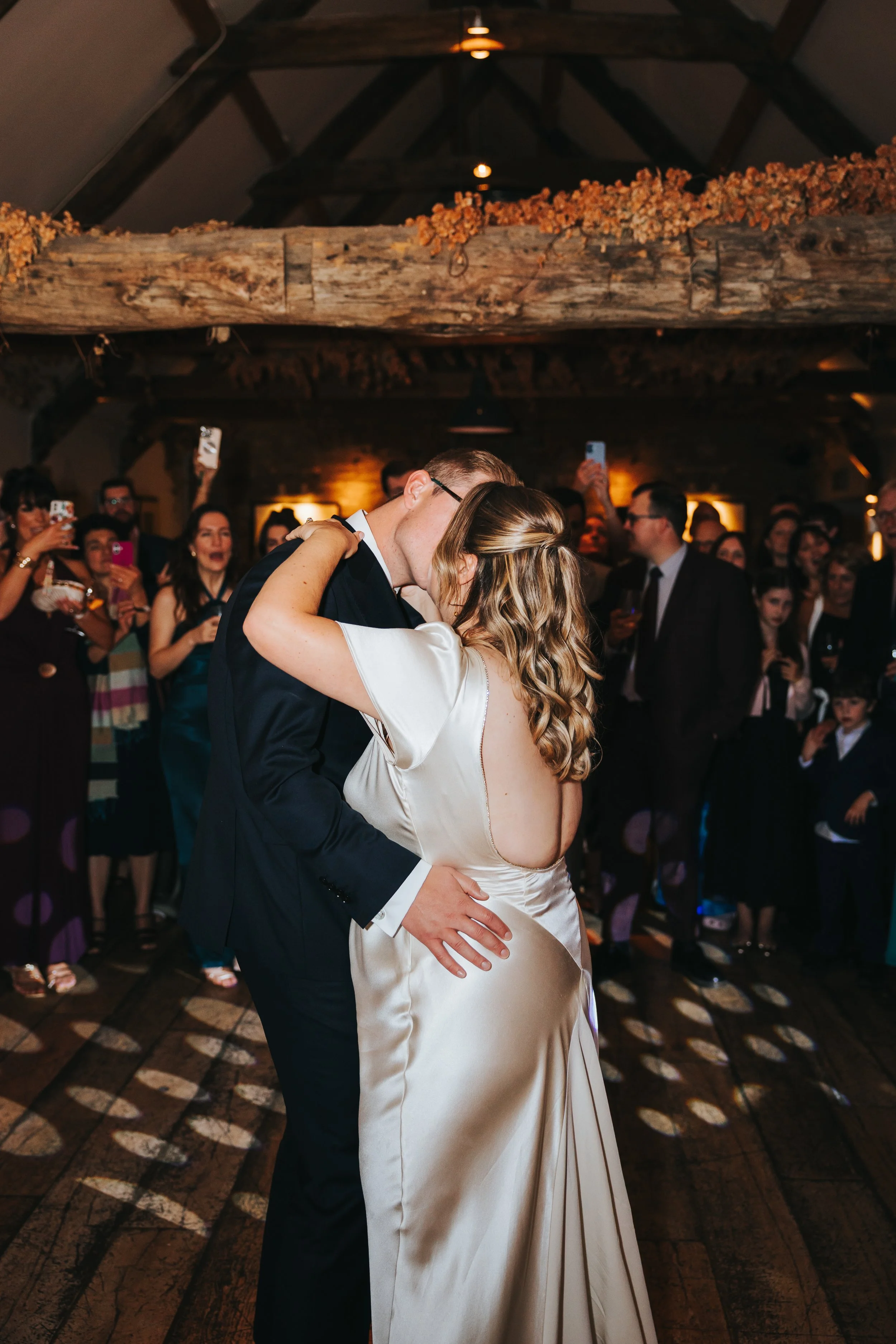 A bride and groom share a first dance at their wedding reception, surrounded by guests taking photos, in a rustic venue with wooden beams and warm lighting.