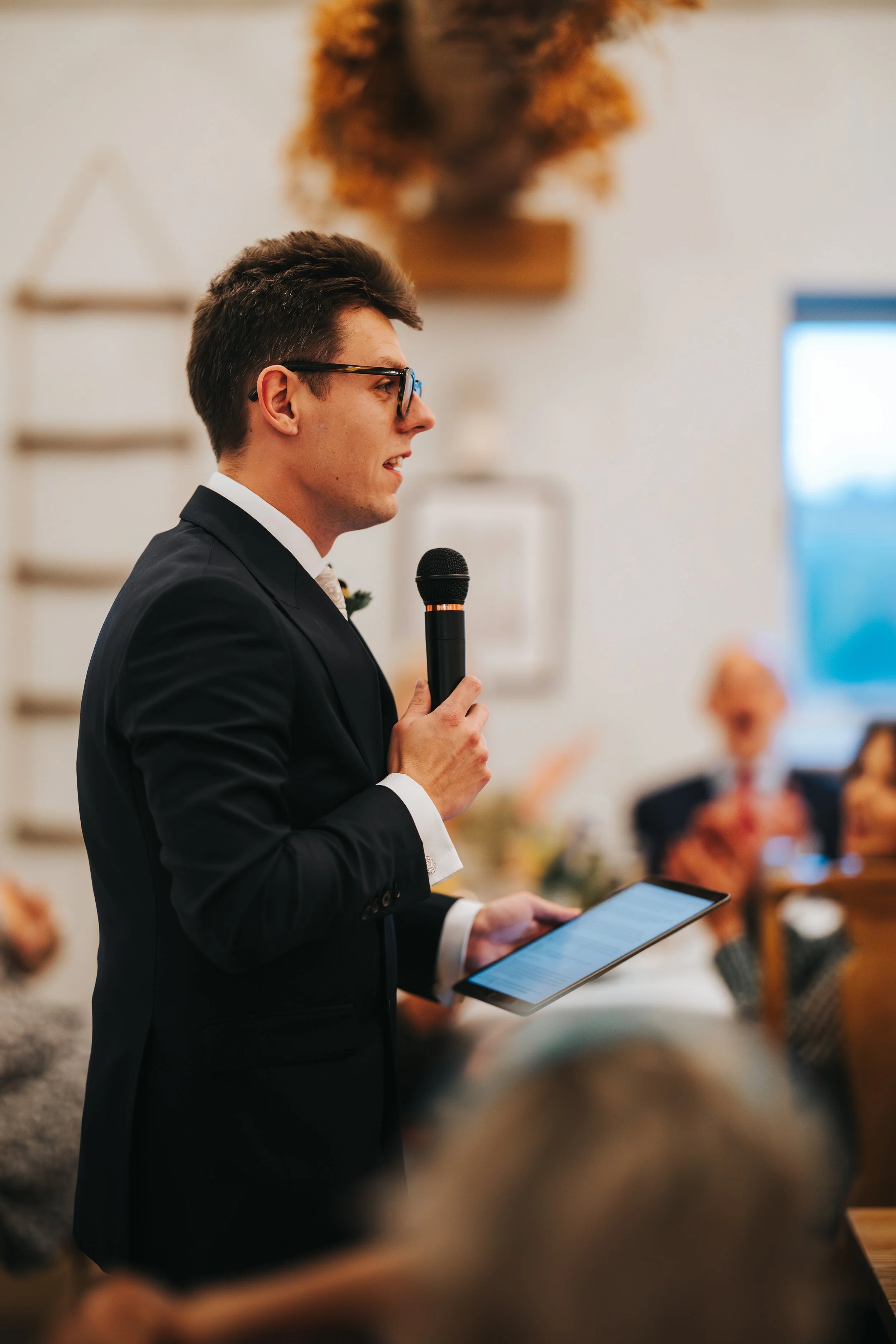 A young man in a black suit and glasses holding a microphone and a tablet, speaking at a formal event or ceremony, with blurred guests in the background.