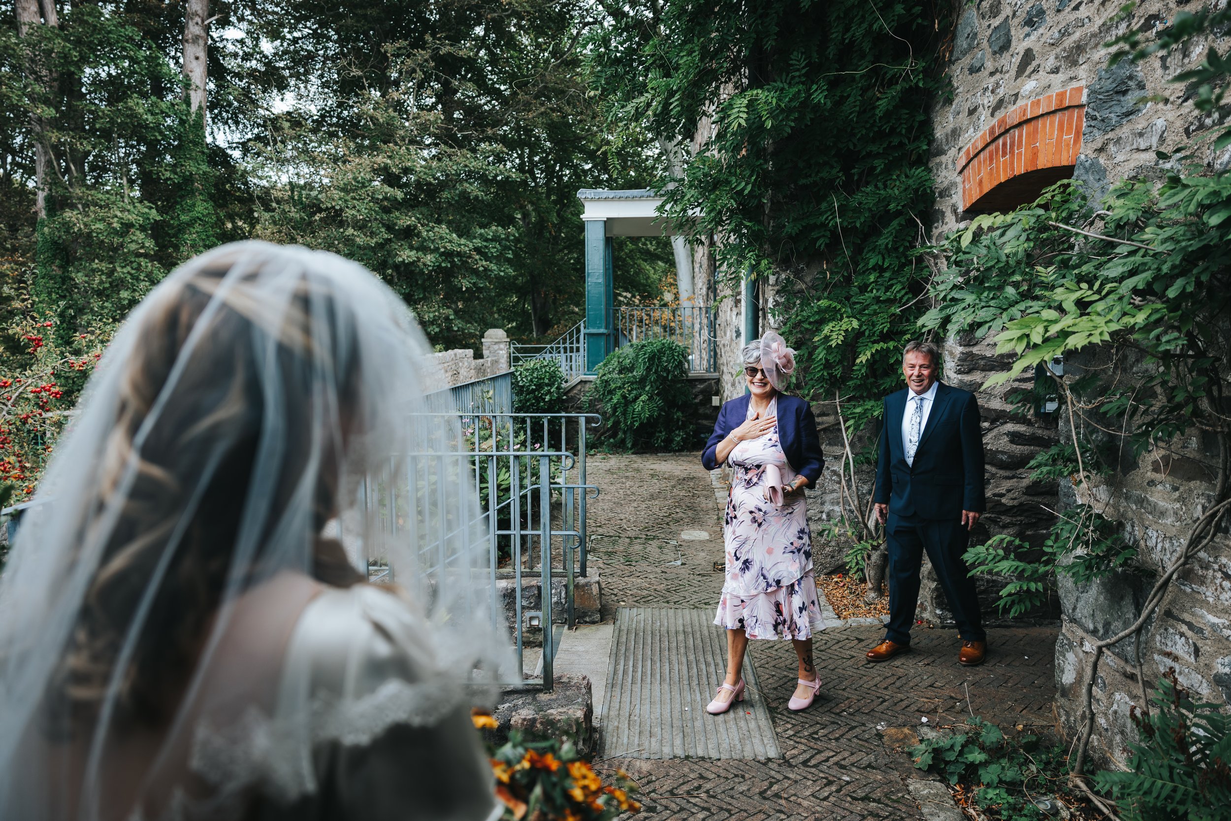 A bride with a veil holds a bouquet of flowers, and two well-dressed guests, a woman in a floral dress with a fascinator and sunglasses, and a man in a dark suit, smile and laugh as they greet her outdoors near a stone wall covered in greenery.
