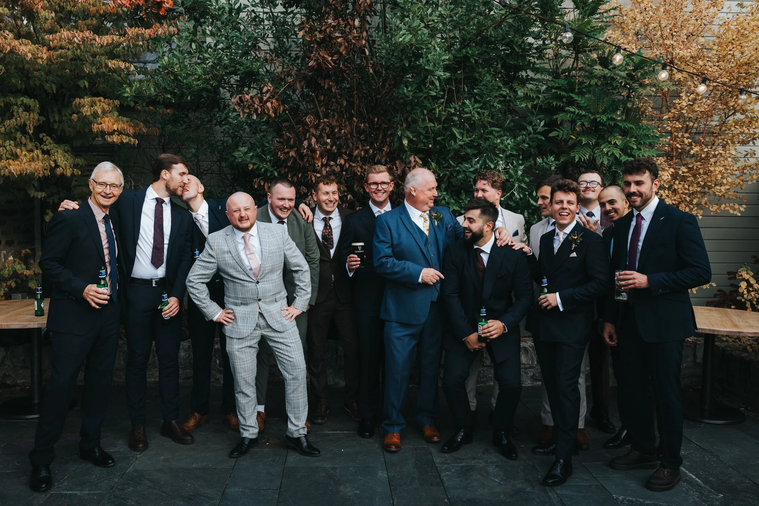 Group of men dressed in suits at a wedding celebration, standing outdoors with trees and string lights in the background, sharing smiles and drinks.