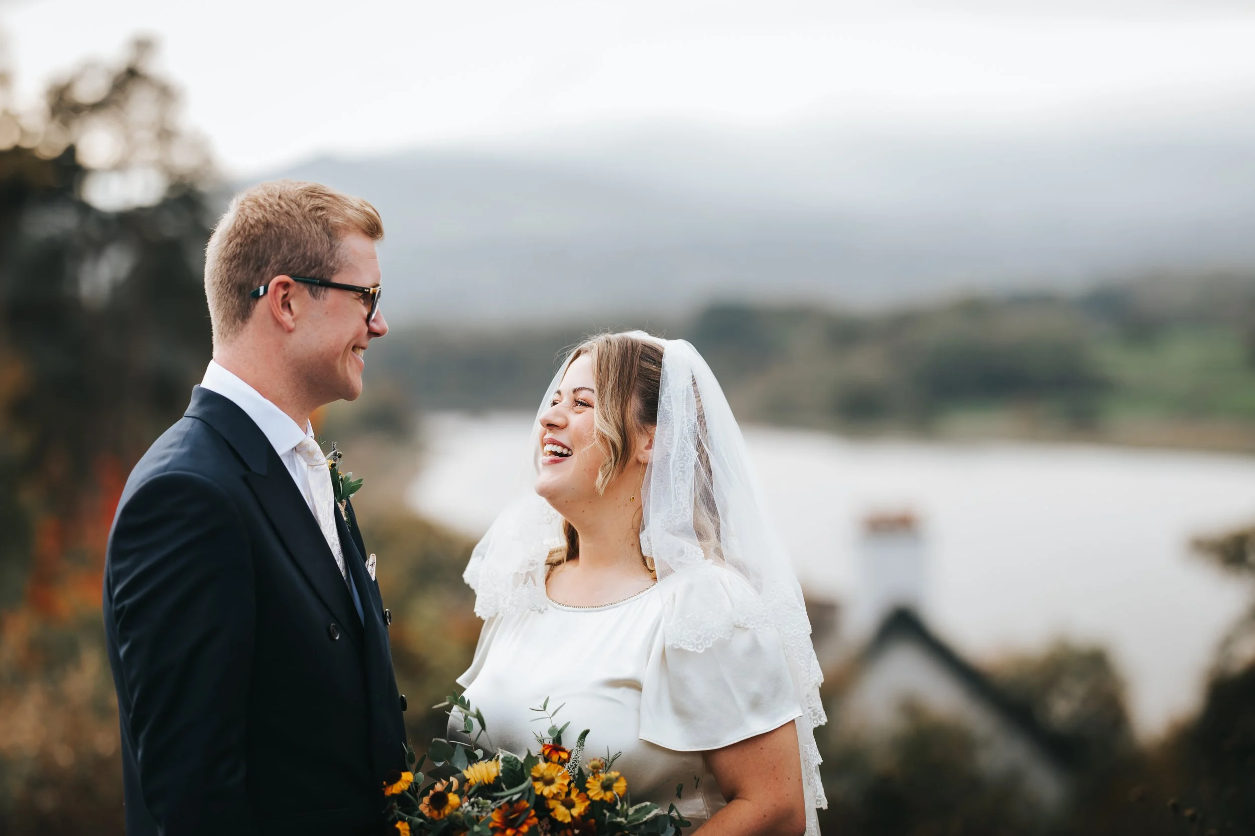 A bride and groom standing outdoors, smiling at each other, with a scenic landscape in the background.