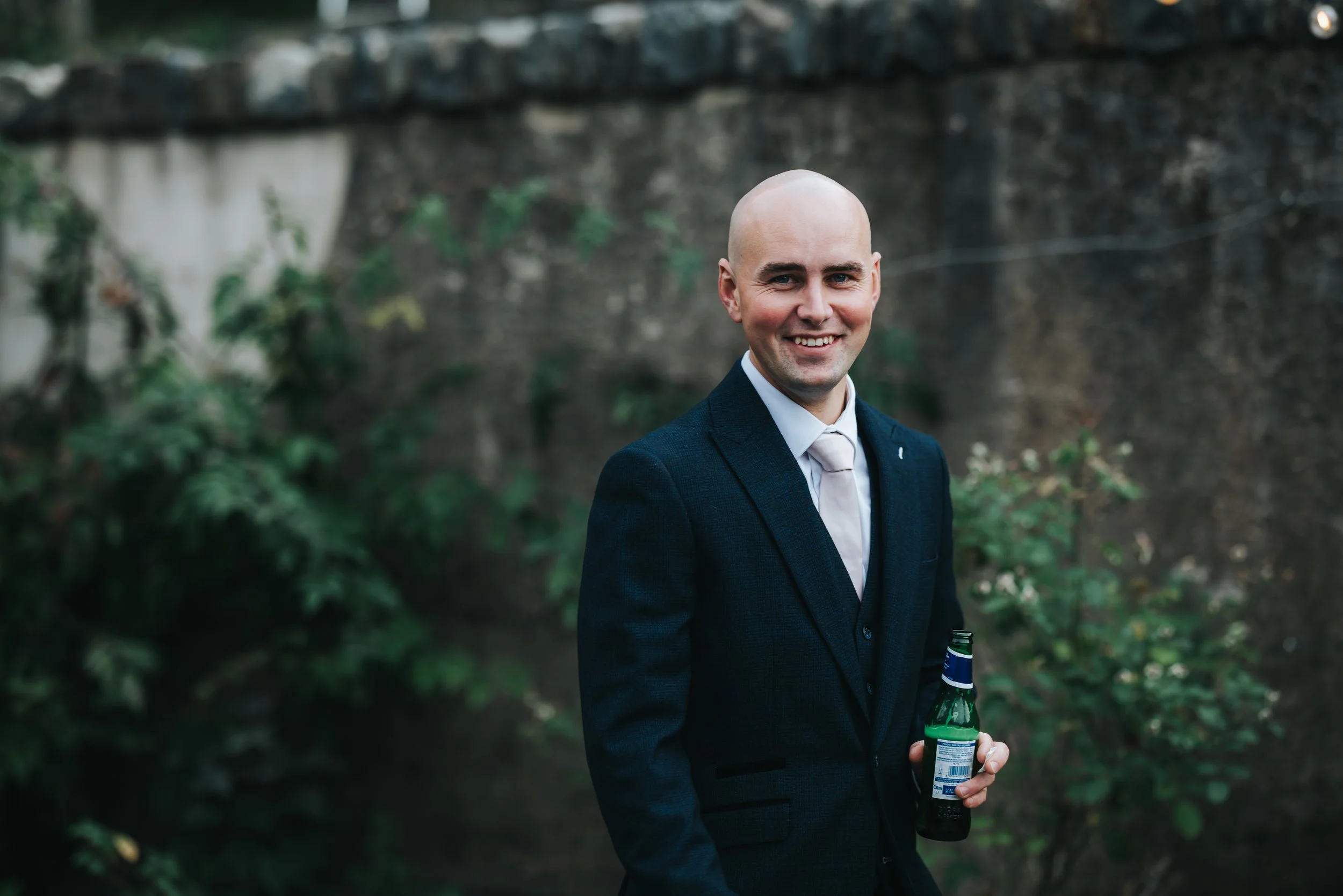 A bald man in a dark suit and light-colored tie standing outdoors, smiling and holding a beer bottle, with greenery and a stone wall in the background.