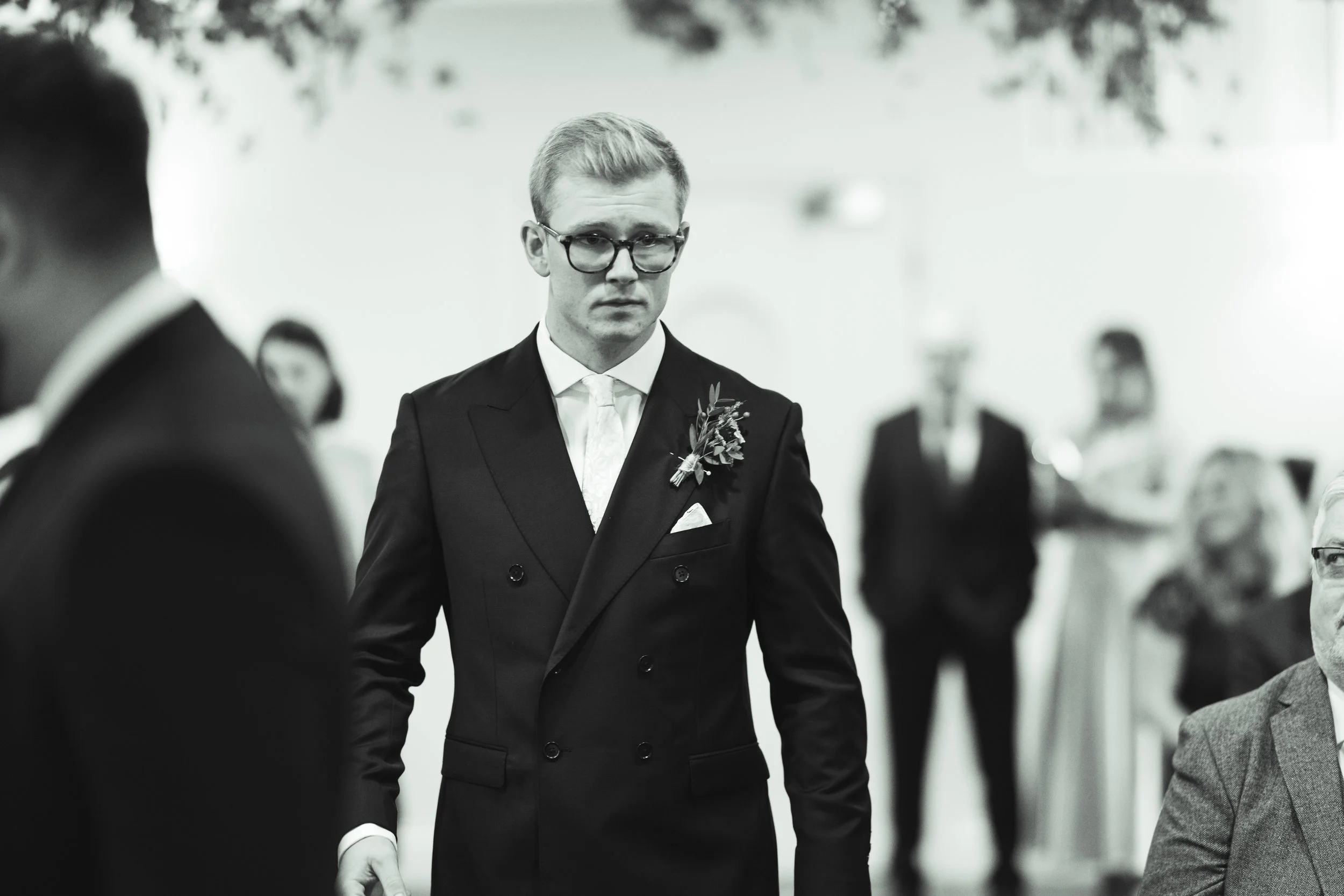A black-and-white photo of a young man in a double-breasted tuxedo with a boutonnière, glasses, and a tie, standing at what appears to be a formal event.