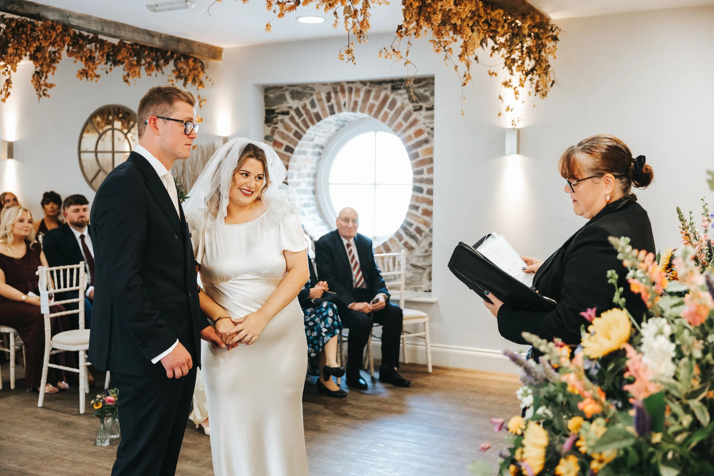 A wedding ceremony with a bride and groom holding hands in front of an officiant, with guests seated and watching in a decorated indoor venue.