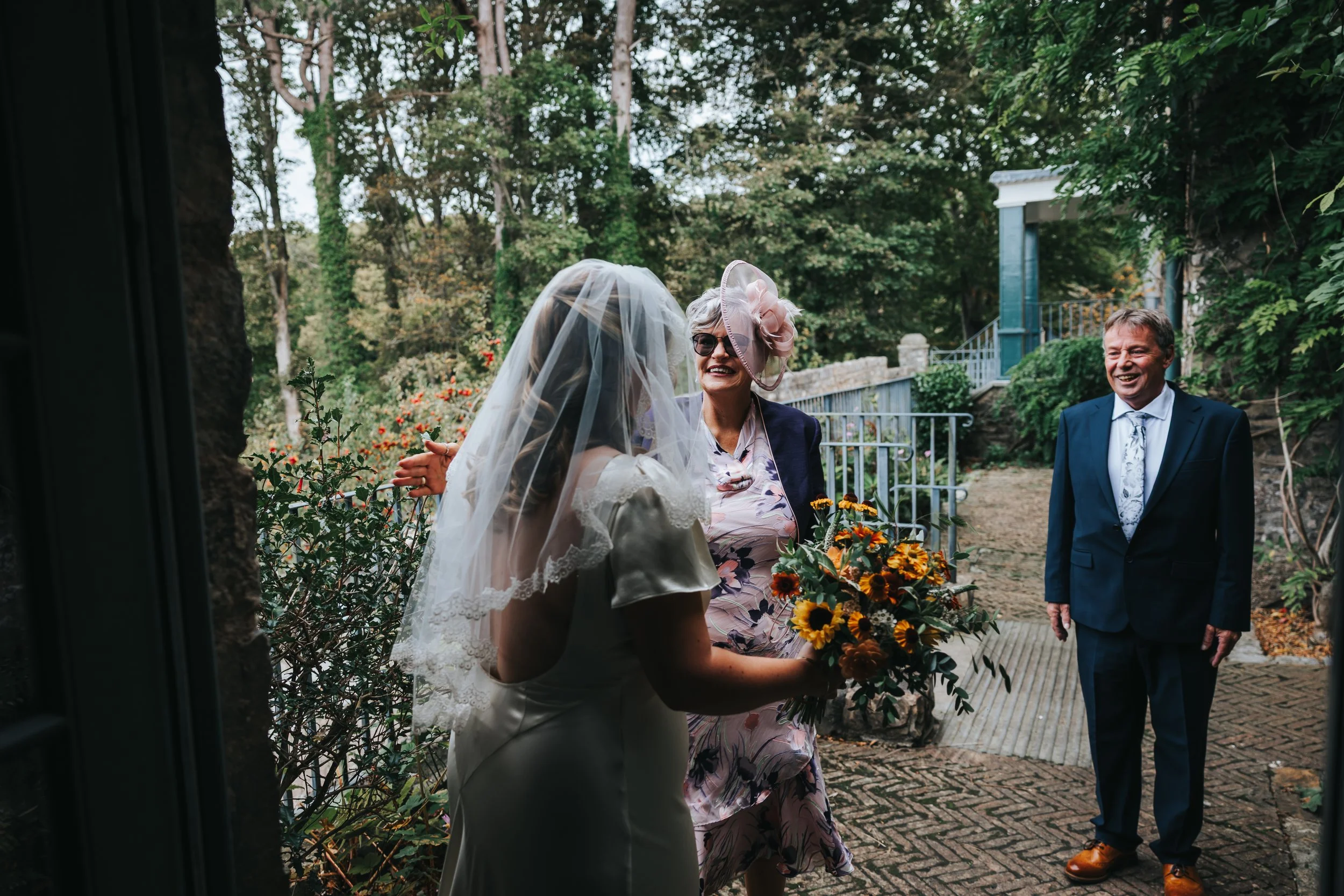 A bride in a wedding dress with a veil and floral lace details is smiling and talking to an older woman wearing a large pink hat, sunglasses, and holding a bouquet of flowers, while a man in a dark suit and tie stands nearby smiling in an outdoor gar