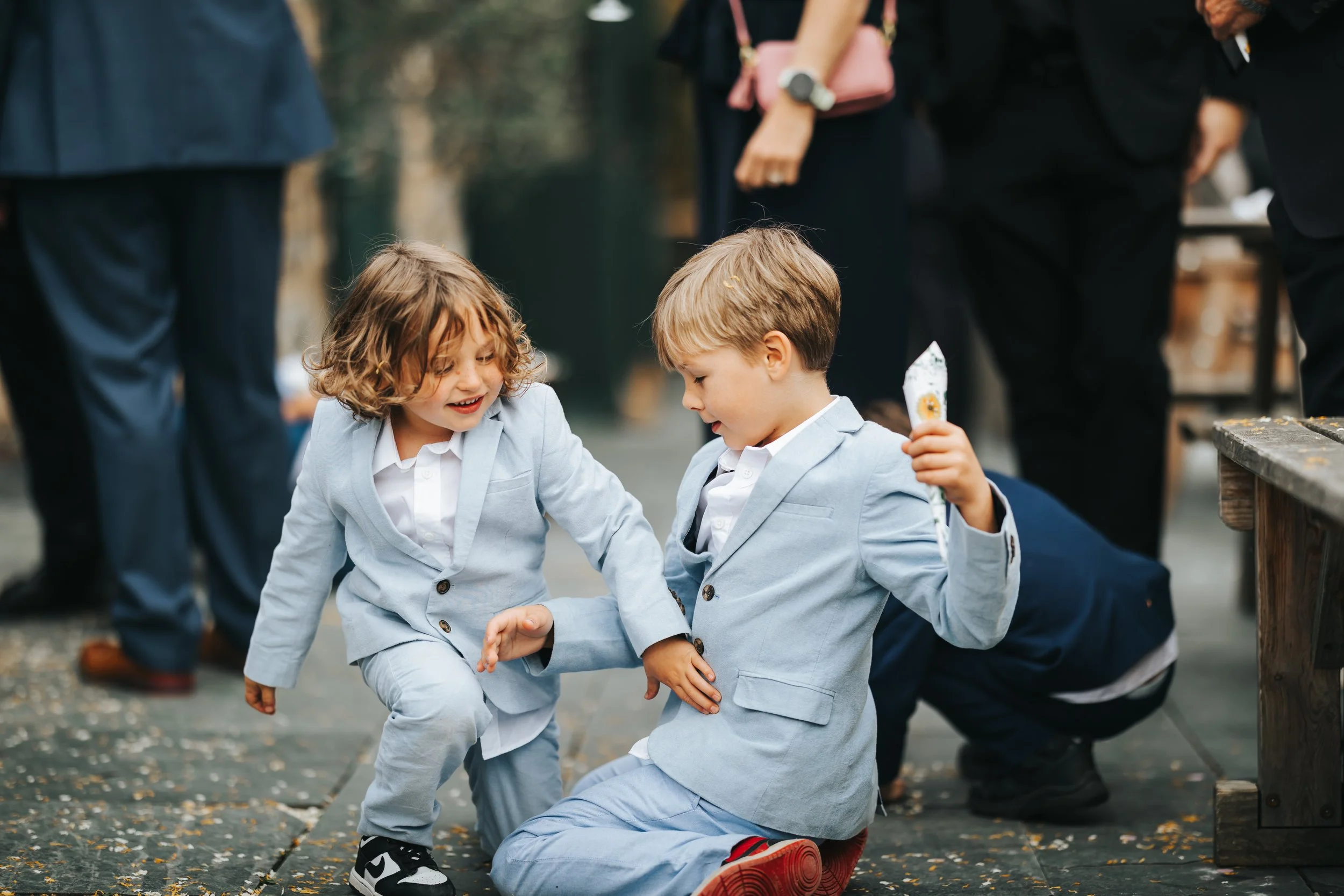 Two young children, a girl and a boy, dressed in light blue suits, are playing together at an outdoor event, with the girl smiling and the boy holding a small snack.