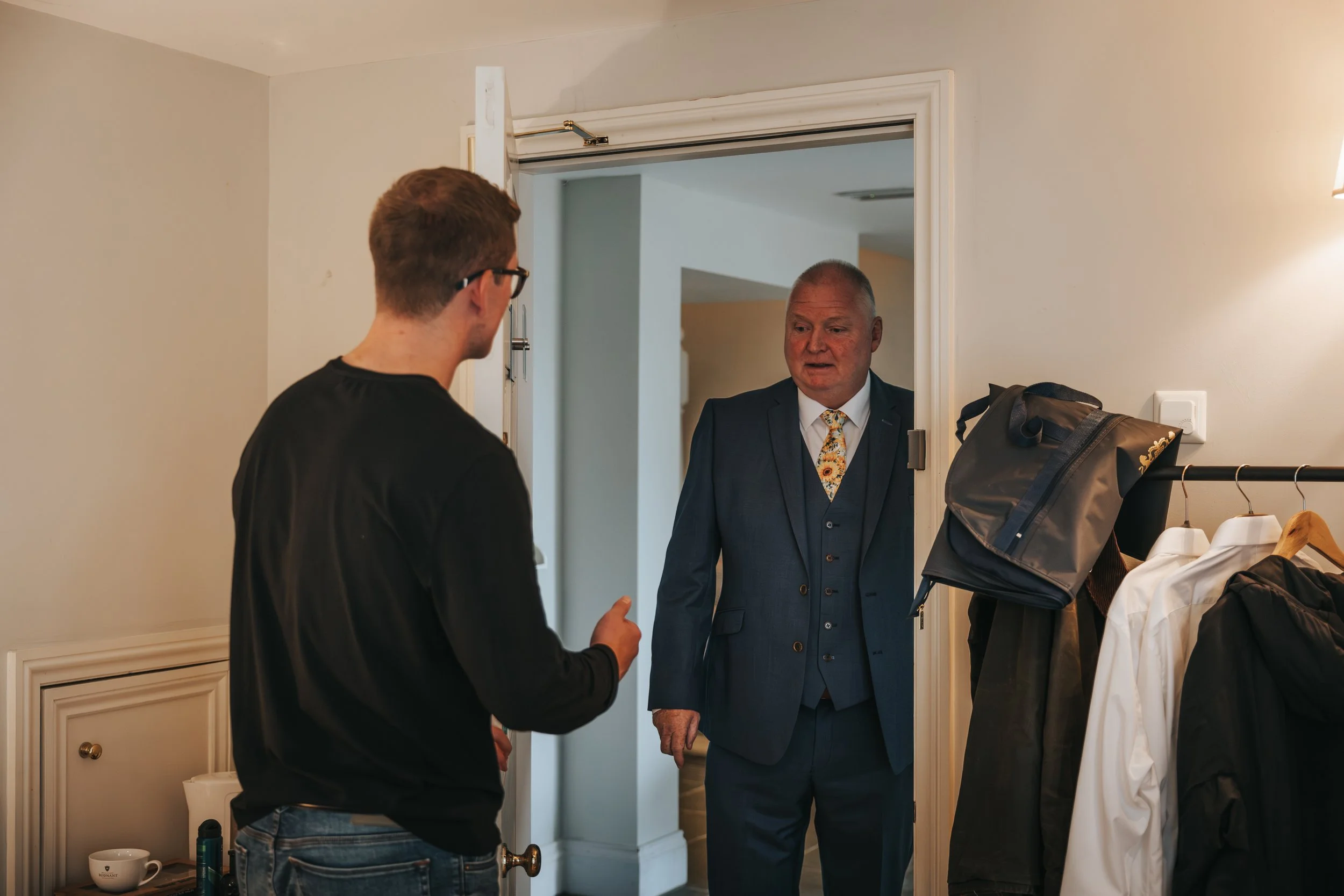 Two men, one in a black shirt and glasses, talking to an older man in a blue suit and patterned tie, standing in a doorway inside a room with a coat rack and a small table.