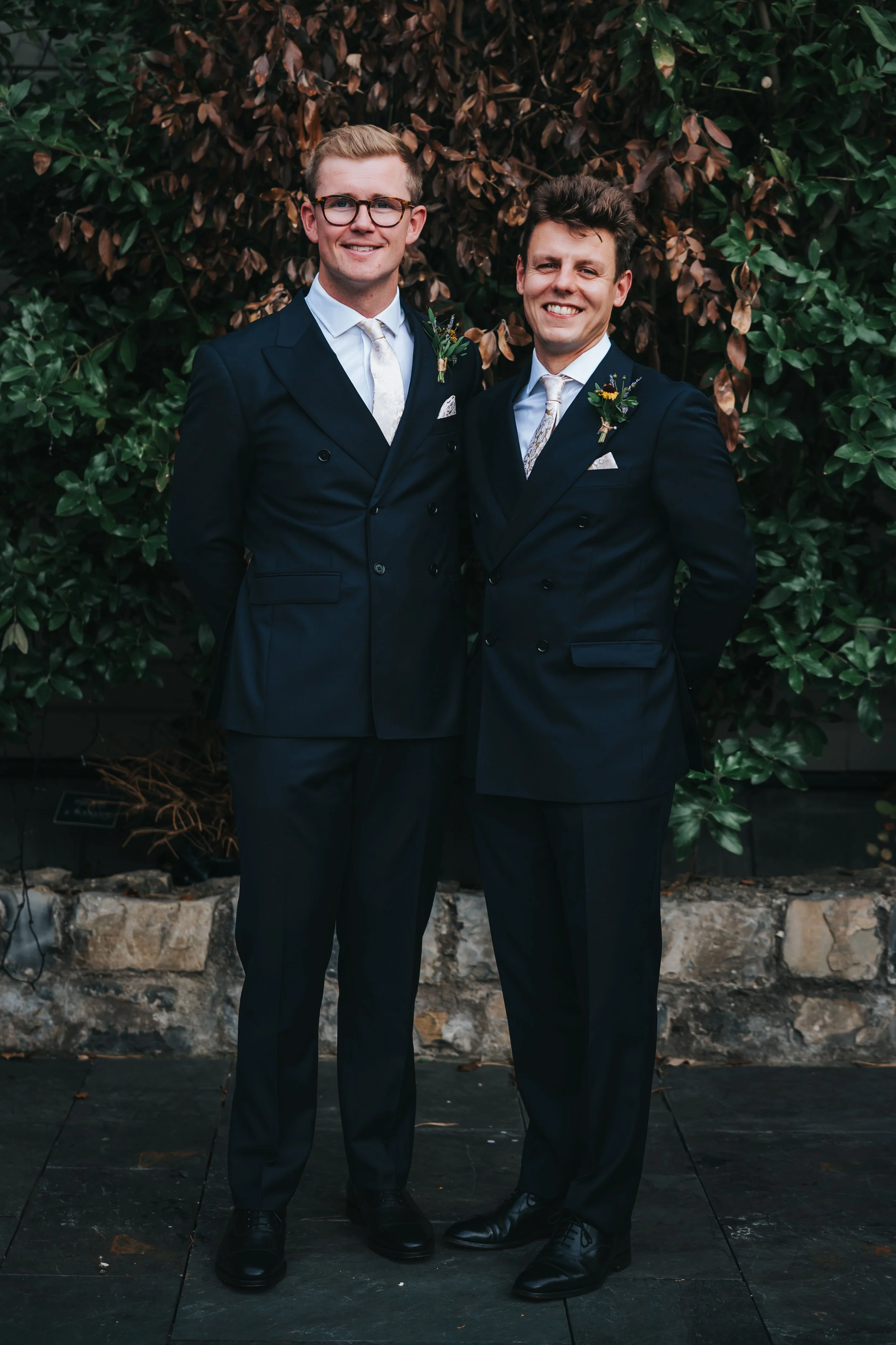 Two men in dark suits stand close together outdoors, smiling, with a backdrop of leafy bushes and stone wall.