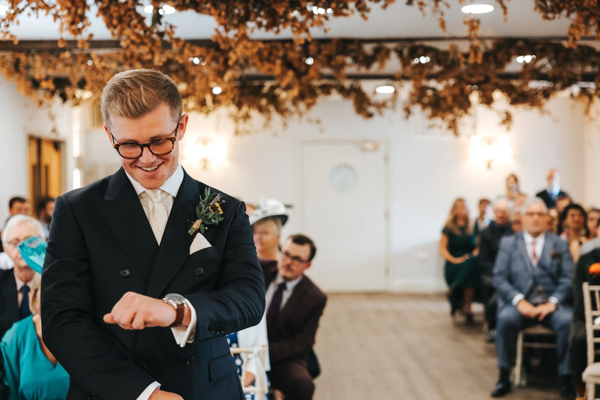 A young man in a black suit with a boutonnière is smiling and looking at his watch during a wedding ceremony. Guests are seated in the background in an indoor venue decorated with autumn-themed hanging ornaments.