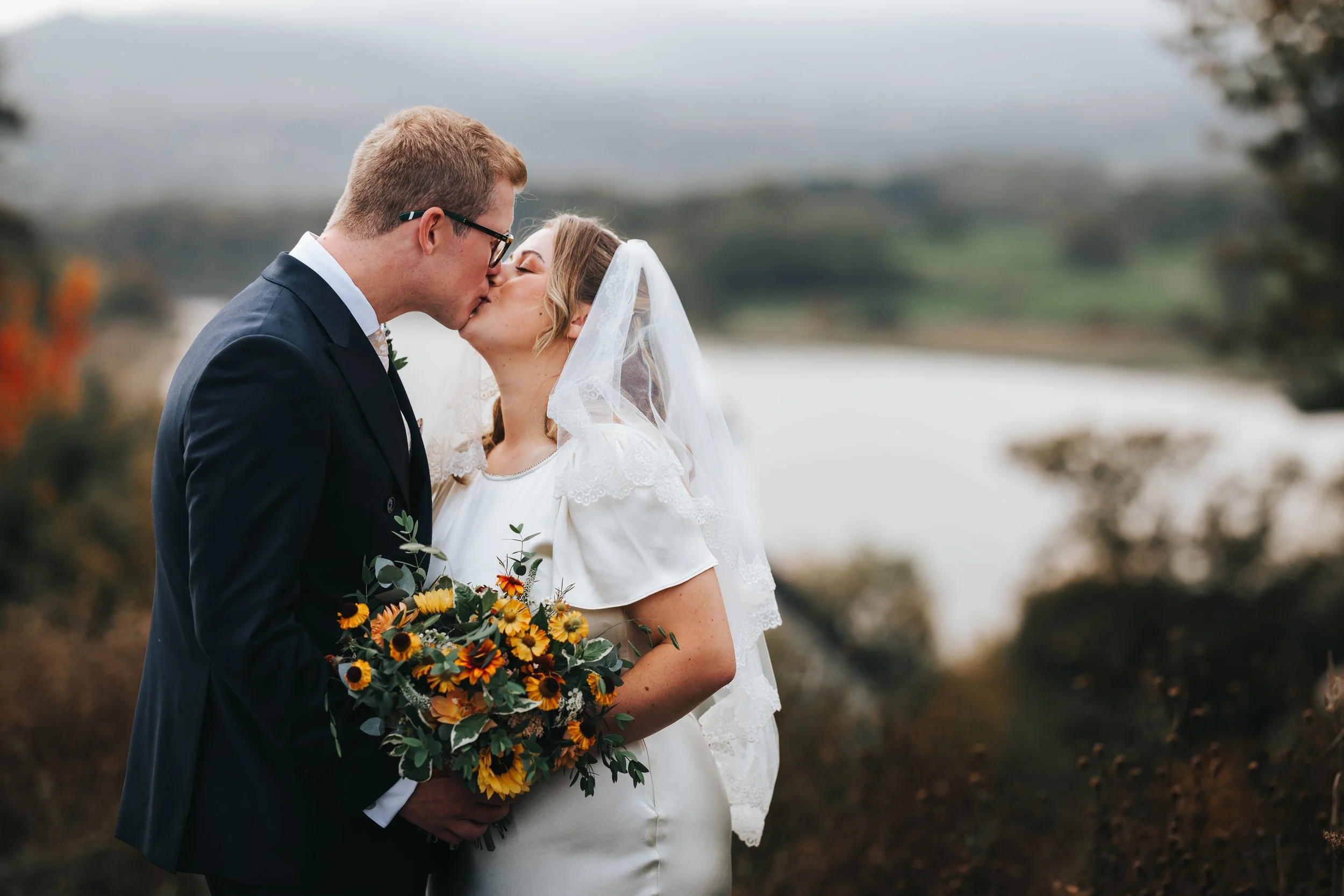 A couple dressed in wedding attire sharing a kiss outdoors, with a scenic background of a lake and trees.