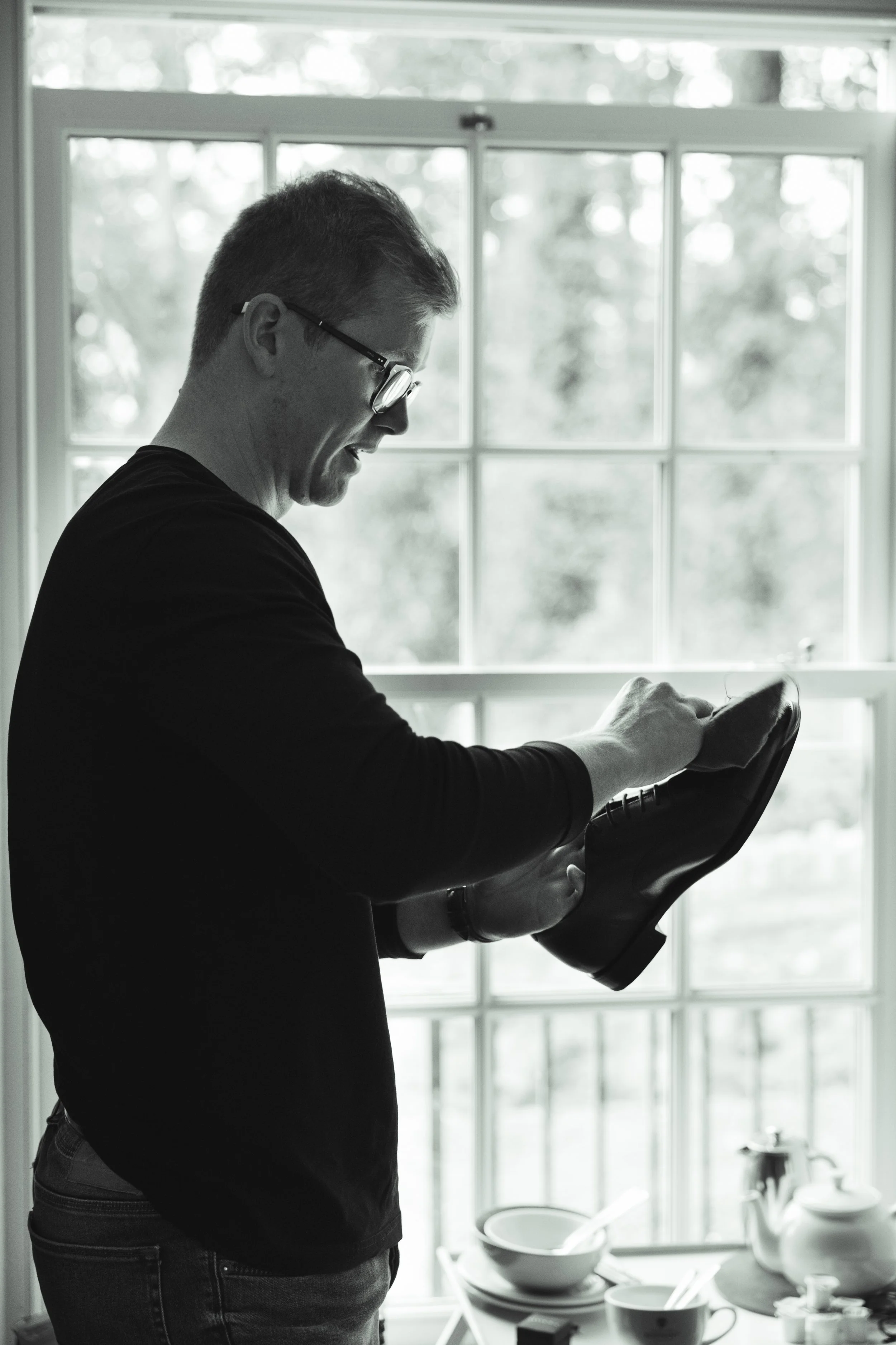A man wearing glasses and a black shirt, smiling and holding a shoe, standing in front of a large window with a view of trees outside.