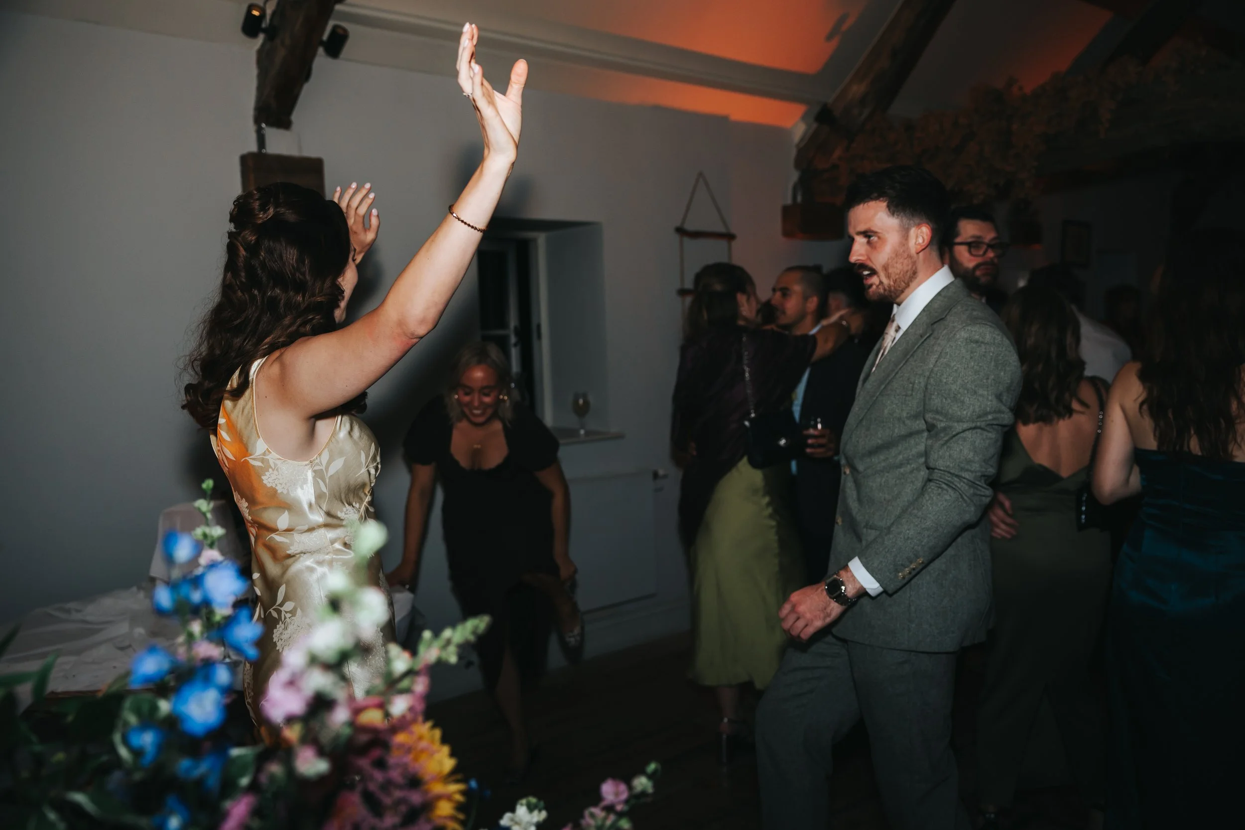 People dancing and socializing at a celebration or party, with a woman raising her arms and a man wearing a suit in the foreground.