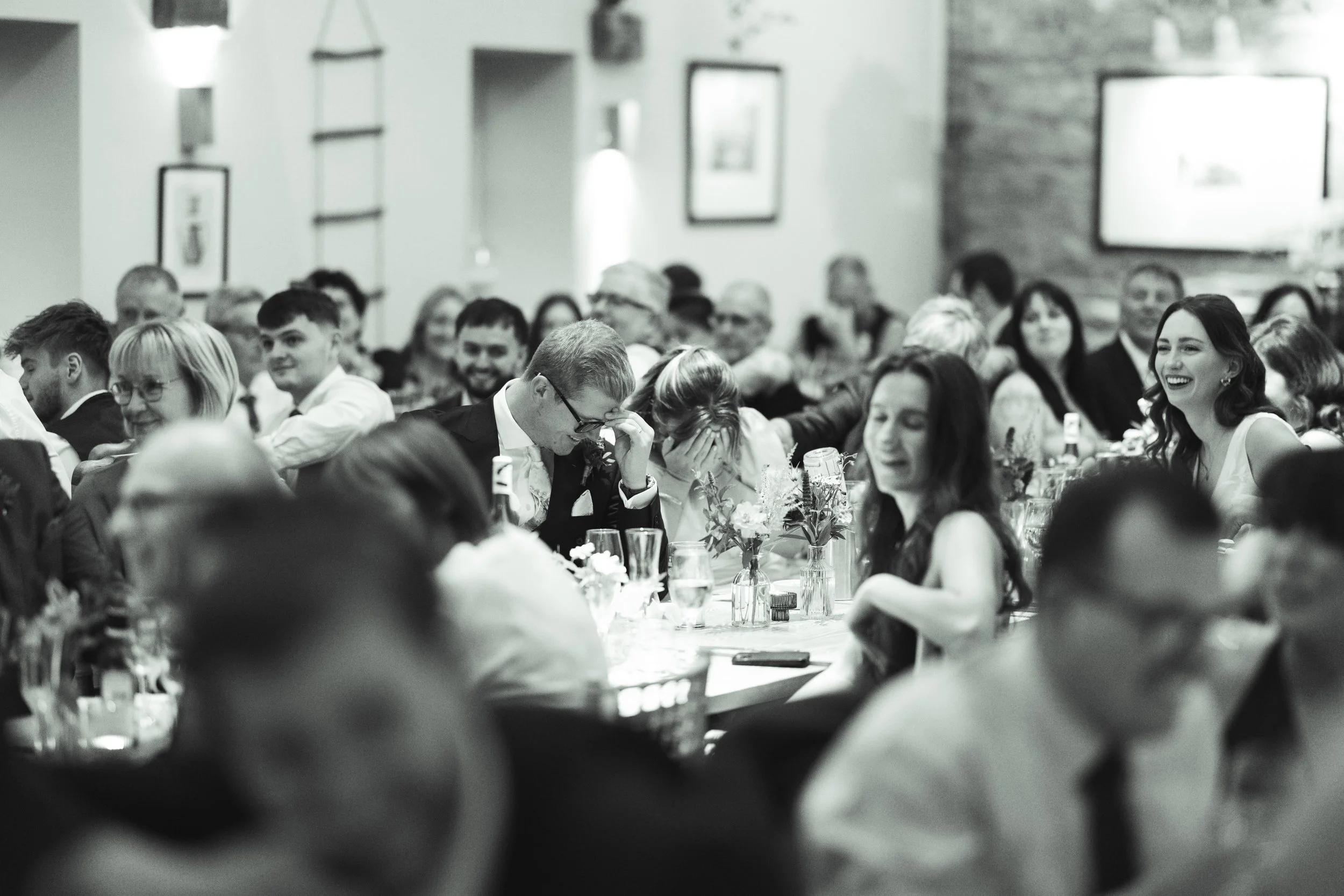 Black and white photo of a group of people sitting at a long table at a formal event, some appear to be laughing or smiling, decorated with flowers and drinks.