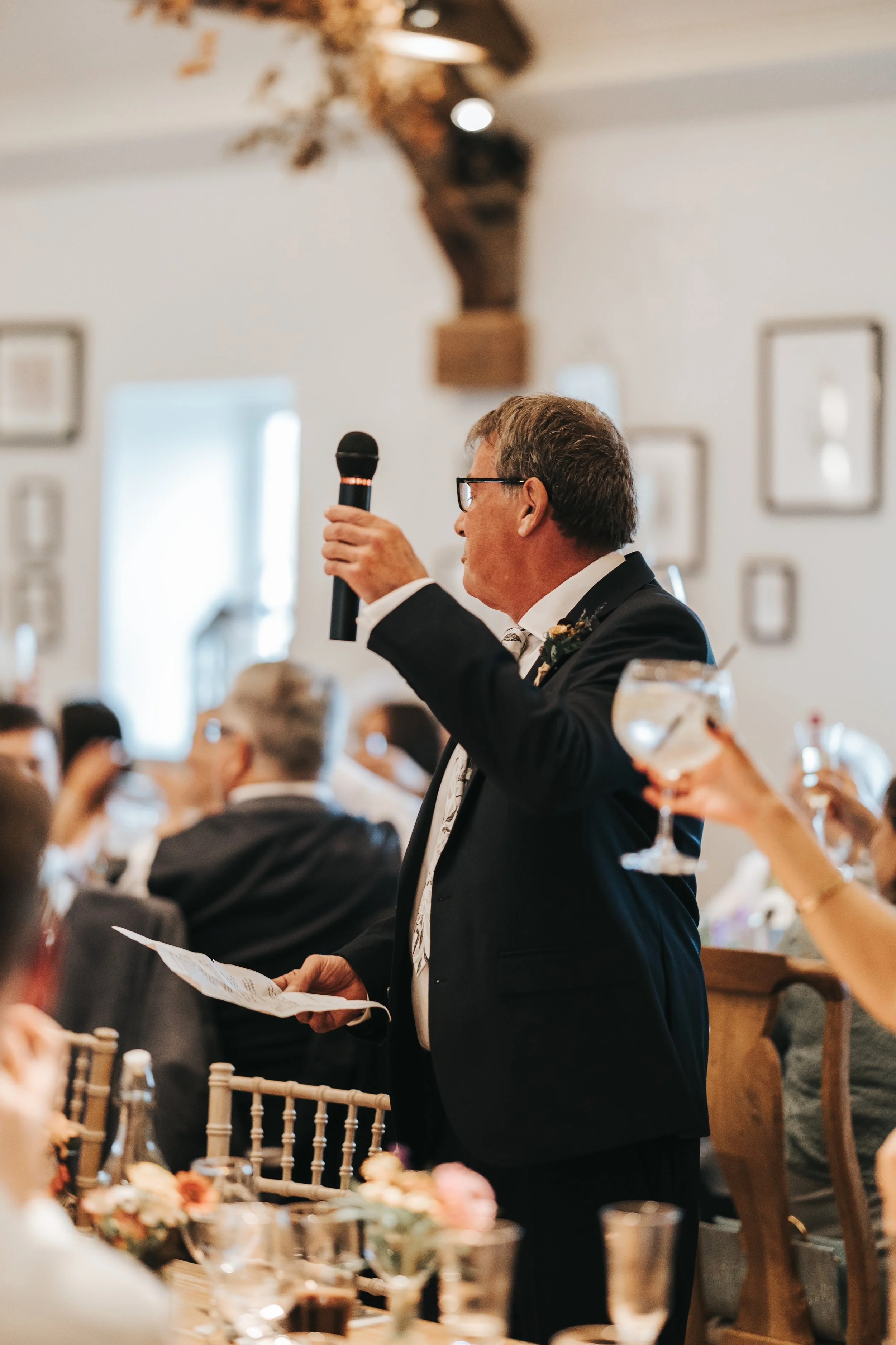 An older man in a black suit and glasses giving a speech at a wedding reception, holding a microphone in one hand and a piece of paper in the other, surrounded by seated guests raising glasses.