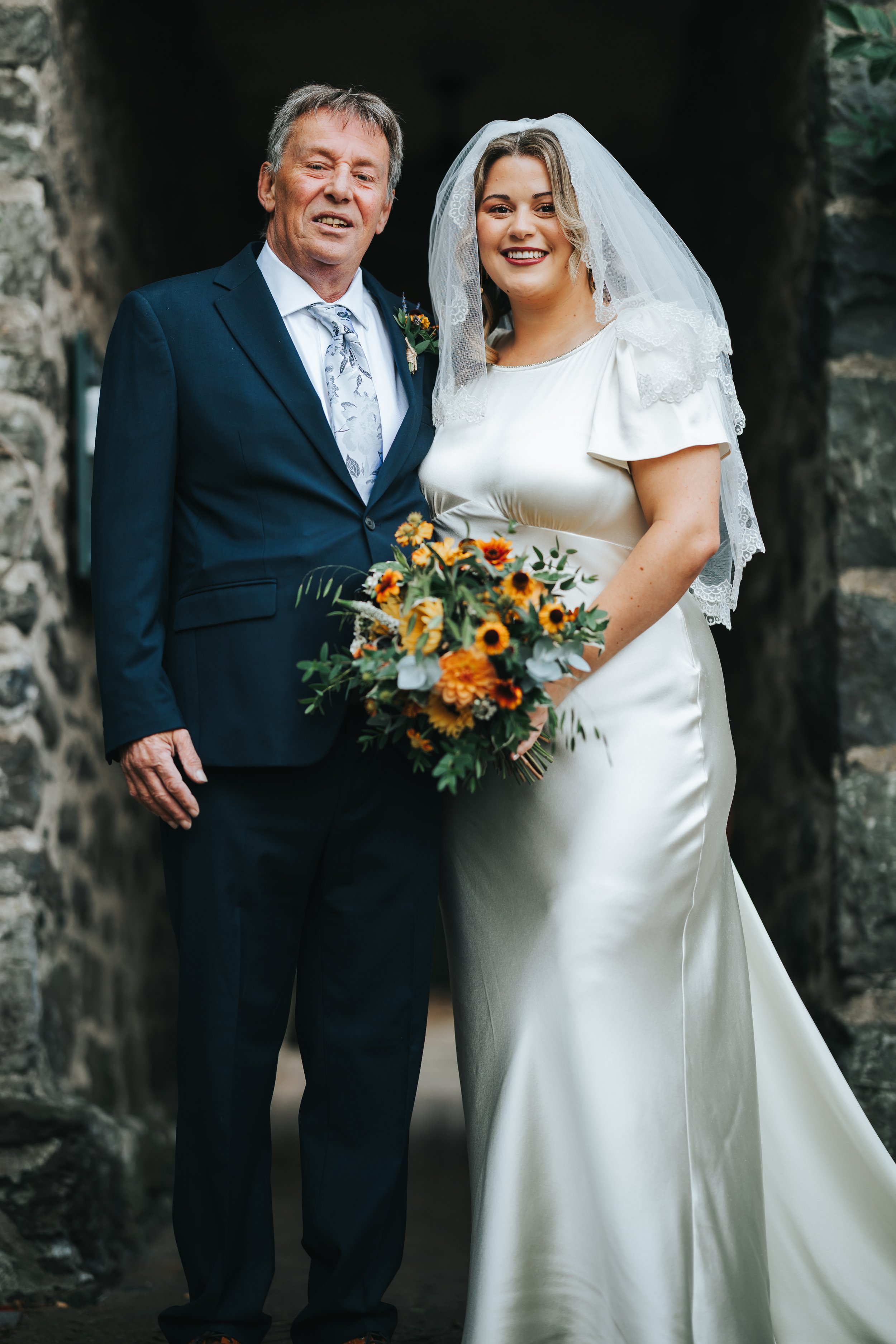A bride and an older man, possibly her father, standing together outdoors near a stone archway. The bride is wearing a white wedding gown and veil, holding a bouquet of orange, yellow, and green flowers. The older man is dressed in a dark suit with a