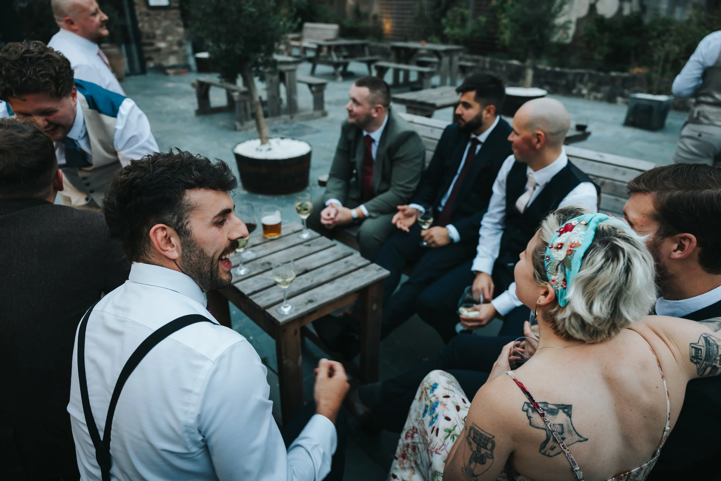 Group of friends enjoying drinks and conversation on a patio, dressed in semi-formal attire, with some tattooed and wearing vintage or retro clothes, sitting around a wooden table with drinks, during an outdoor gathering.