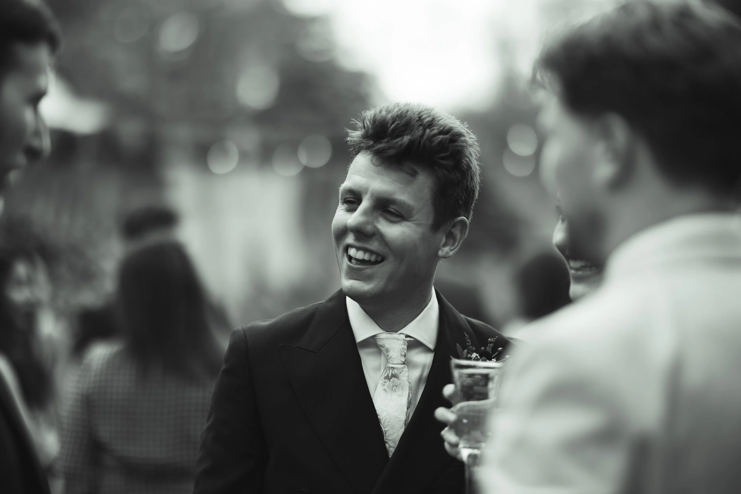 Black and white photo of a smiling young man in a suit at a social gathering, holding a drink, engaging with others.