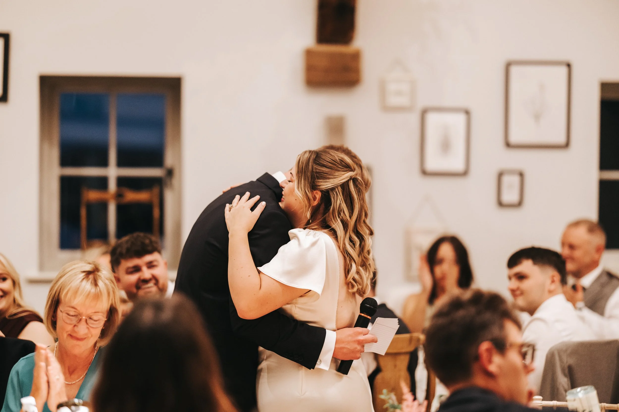 A woman in a white dress hugging a man in a dark suit at a wedding reception, while holding a microphone and notes. Several guests are seated at tables, smiling and clapping.