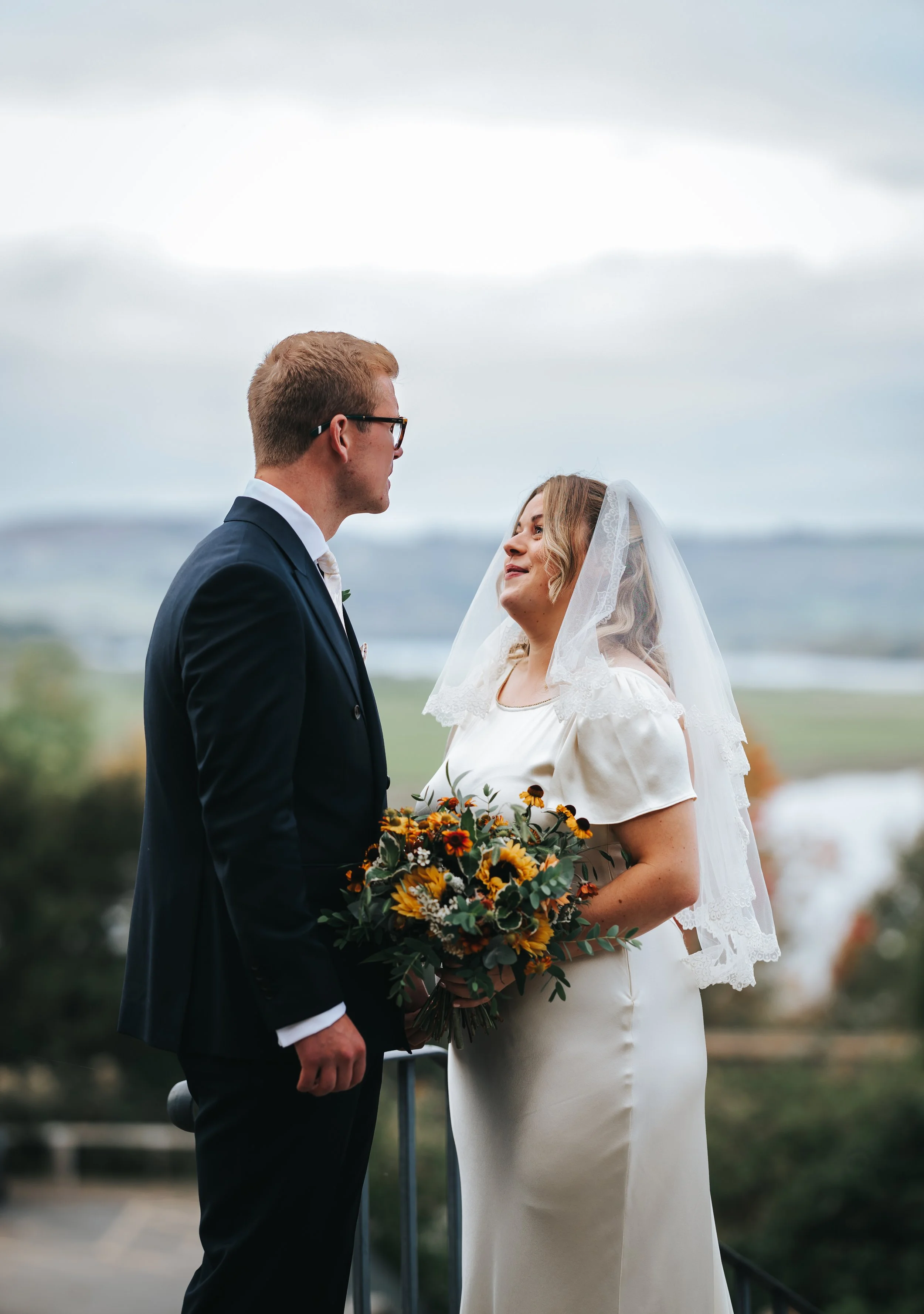 A bride and groom standing facing each other outdoors with a scenic landscape background, the bride holding a bouquet of sunflowers and greenery, dressed in a white gown with a veil, and the groom in a dark suit with glasses.
