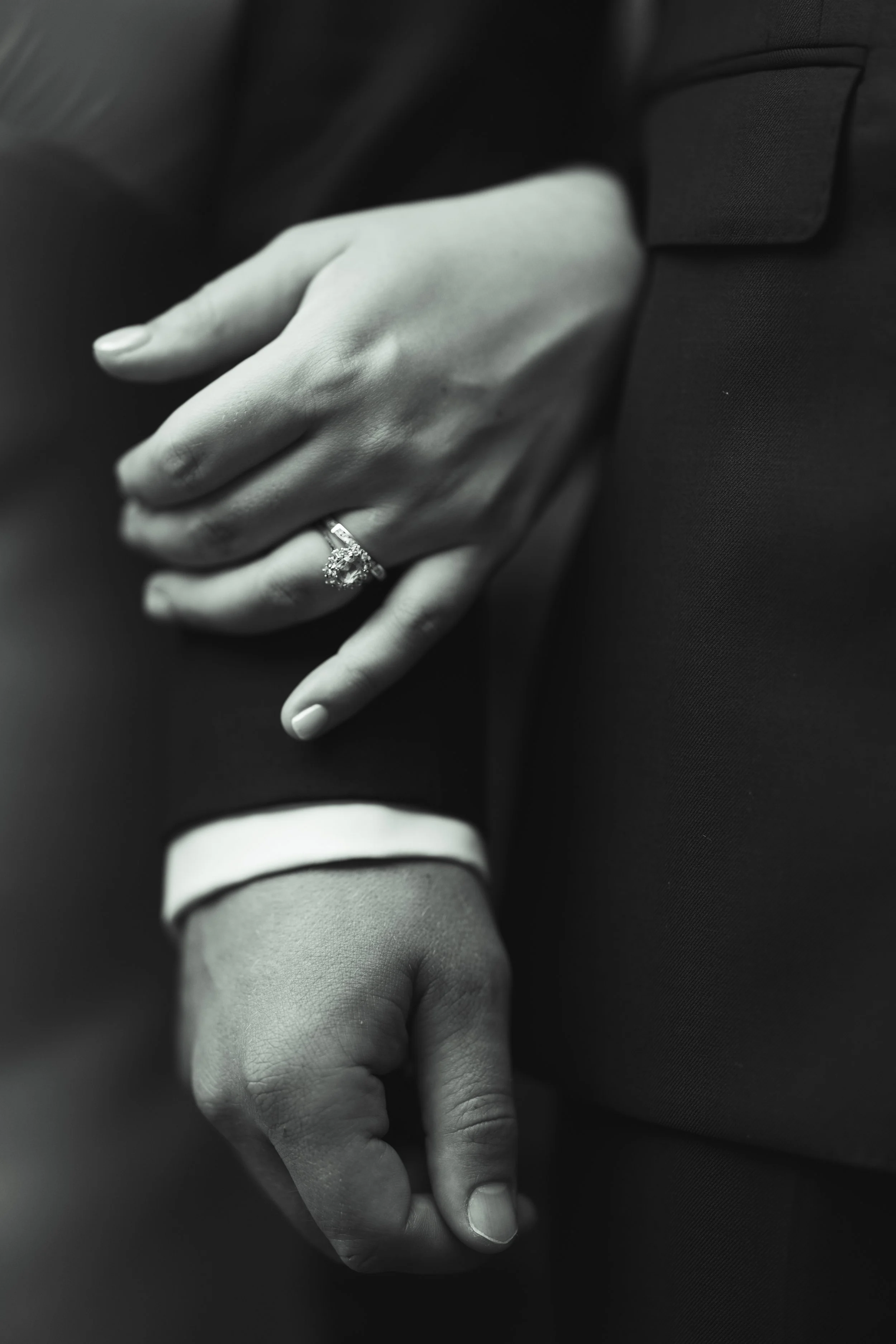 Close-up of a woman's hand with an engagement ring resting on a man's arm, both in formal attire.